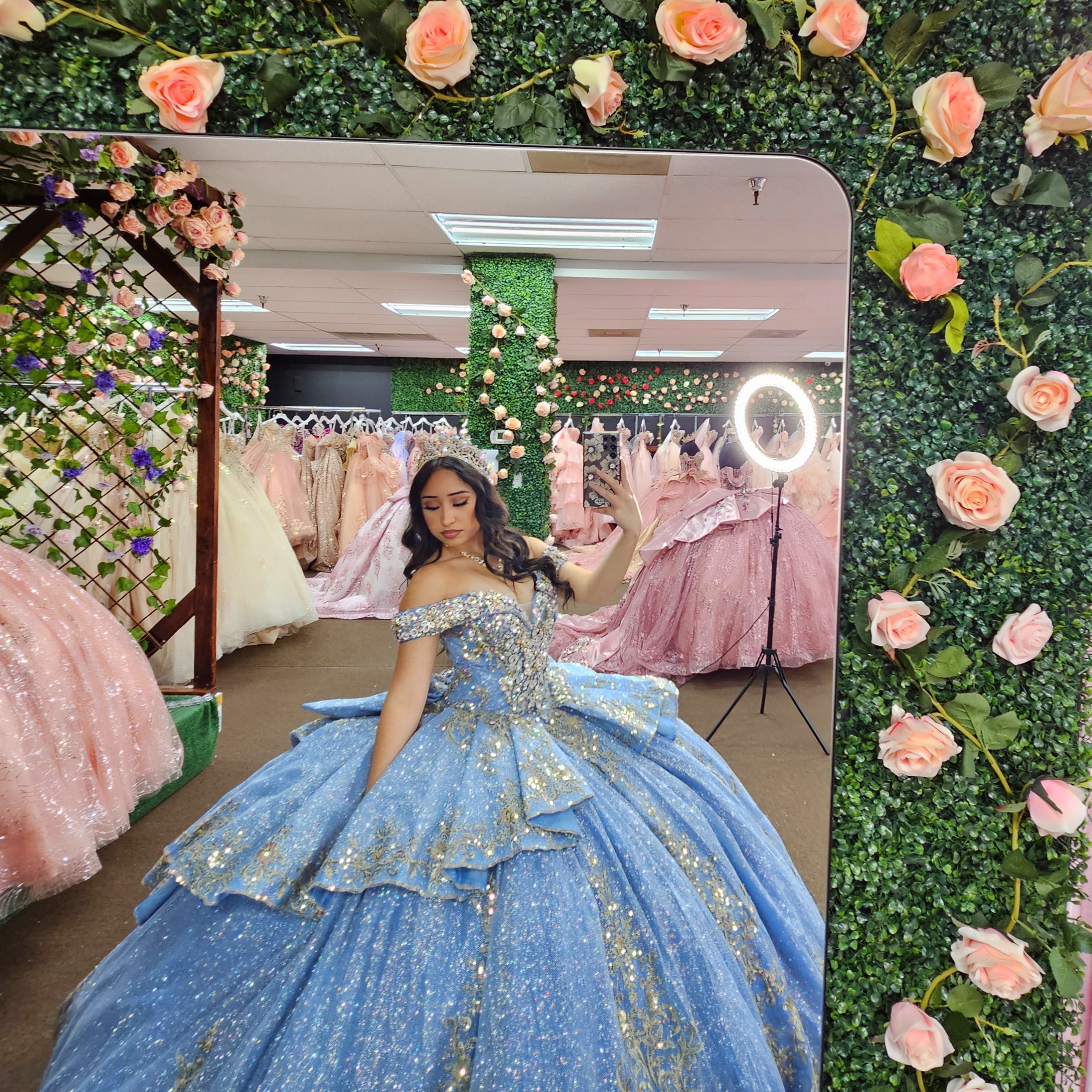 Woman trying on a blue ball gown with gold embroidery in a bridal shop, surrounded by pink and white dresses, with flower decorations and a ring light in the background.