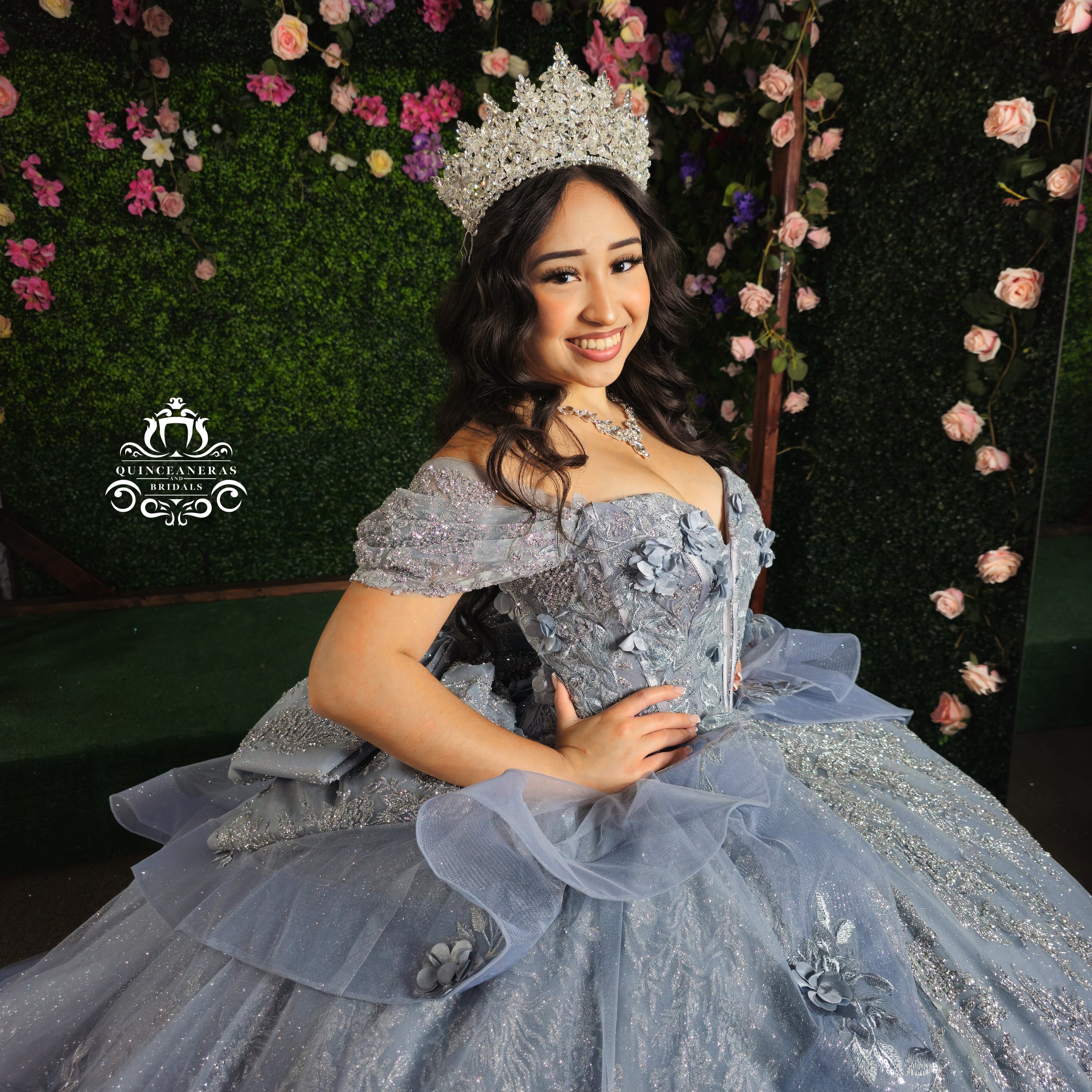 Young woman in a silver gown with floral embellishments, a tiara, and jewelry, sitting in front of a floral backdrop.