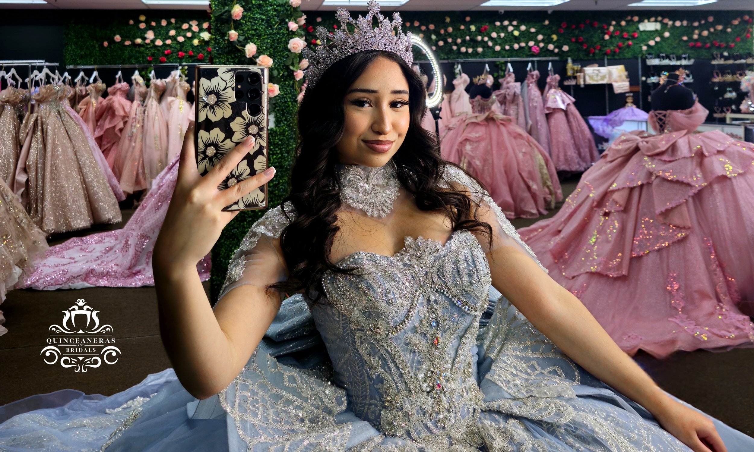 Young woman wearing a silver tiara and an elegant, elaborate silver gown, taking a selfie inside a bridal boutique with pink and gold dresses on display in the background.