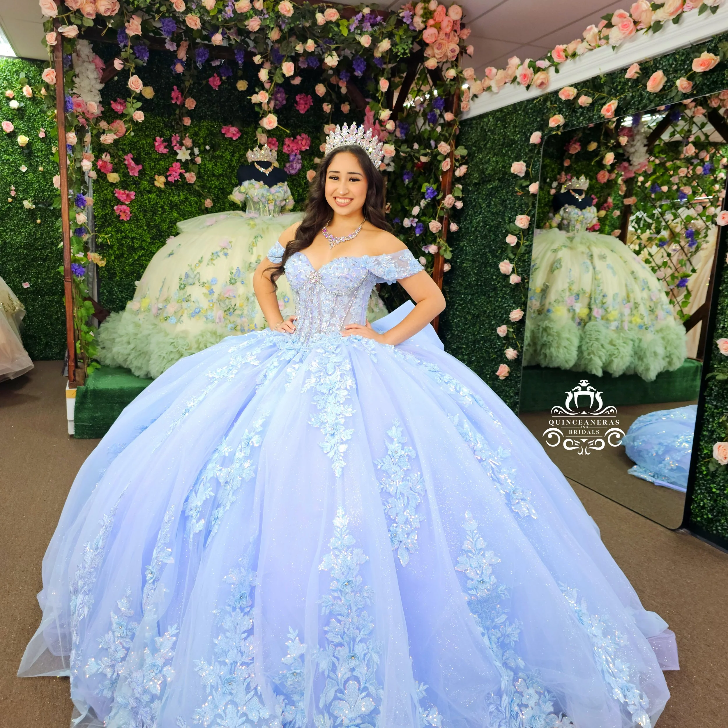 A young woman wearing a light blue ball gown with floral embroidery, standing in front of a decorative floral backdrop with pink and purple flowers, a mirror, and a dress with a matching floral skirt.