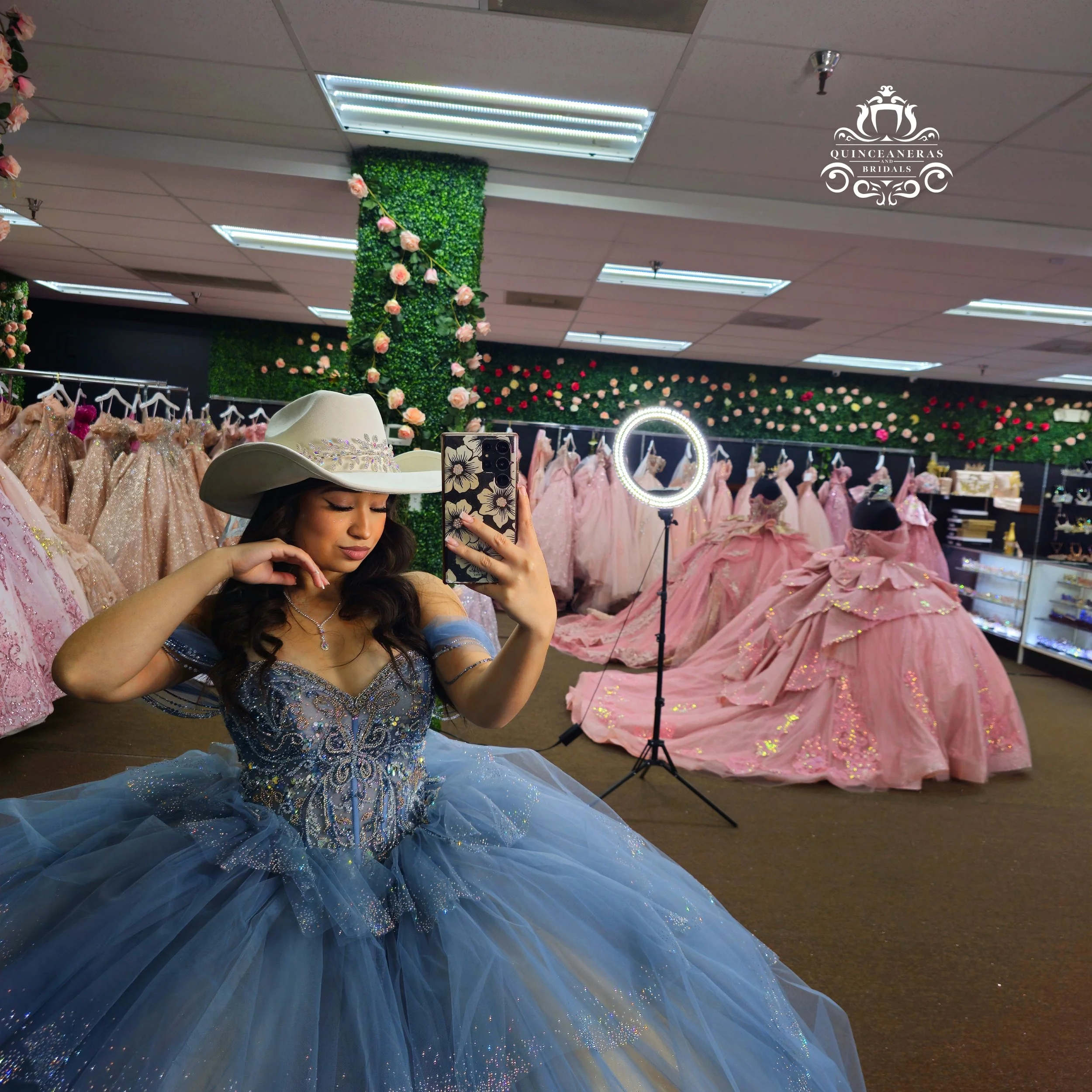 Young woman in a blue quinceañera dress with a white wide-brimmed hat taking a selfie in a dress shop decorated with pink ballgown dresses and floral displays.