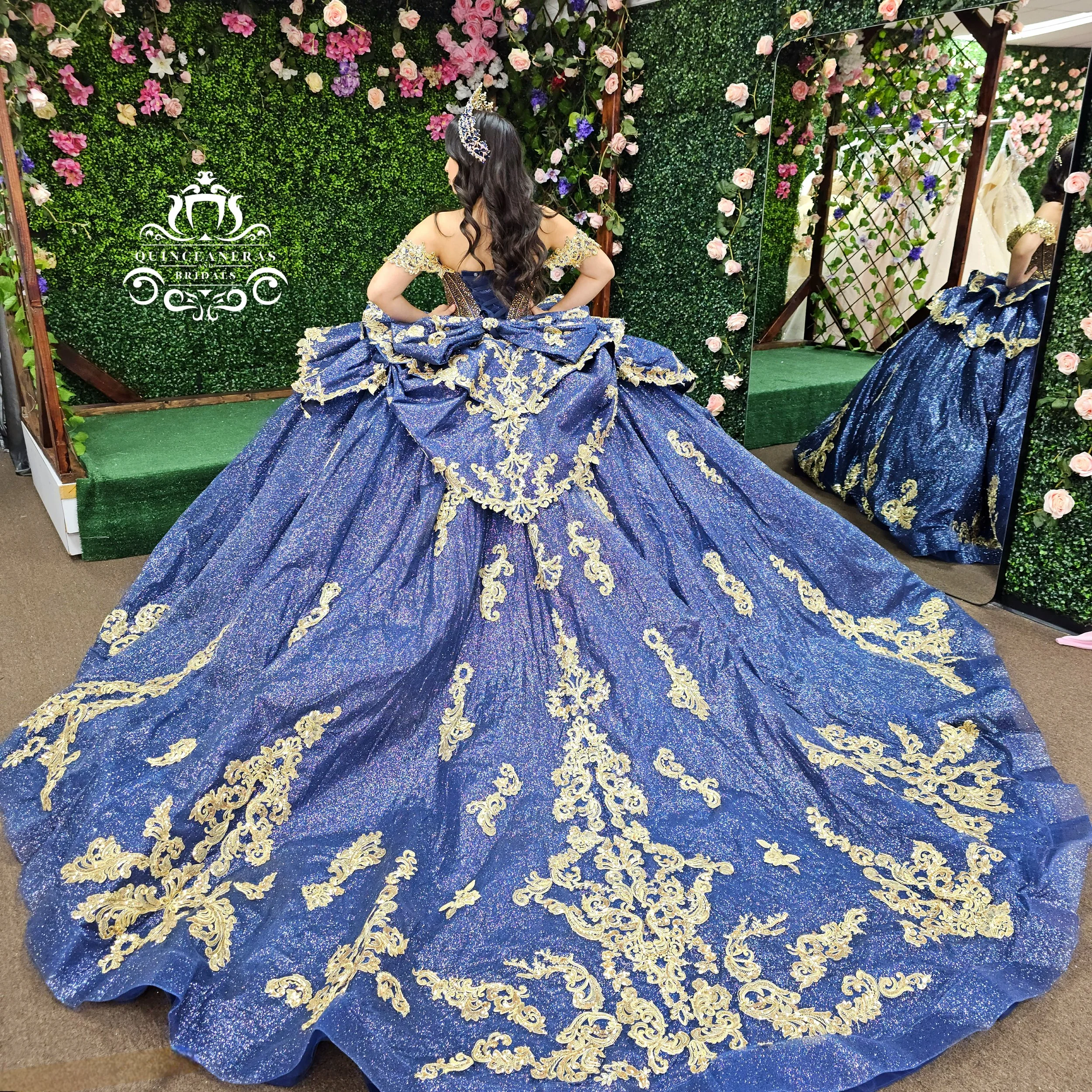 A woman in a royal blue and gold ornate ball gown standing in front of a floral backdrop and mirror, trying on a wedding dress.