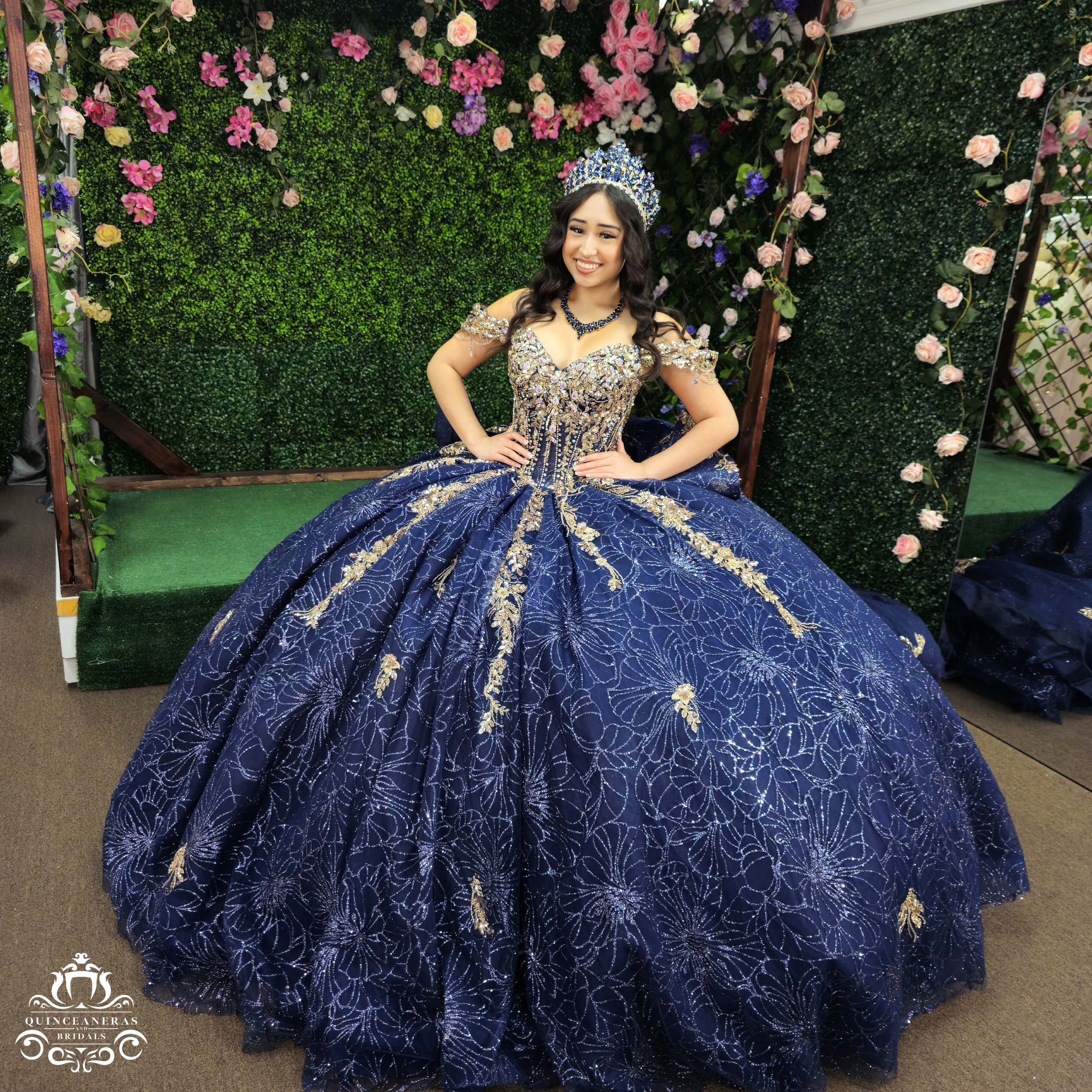 Young woman in a royal blue ball gown with gold embroidery and beading, wearing a matching crown and jewelry, sitting in front of a floral backdrop.