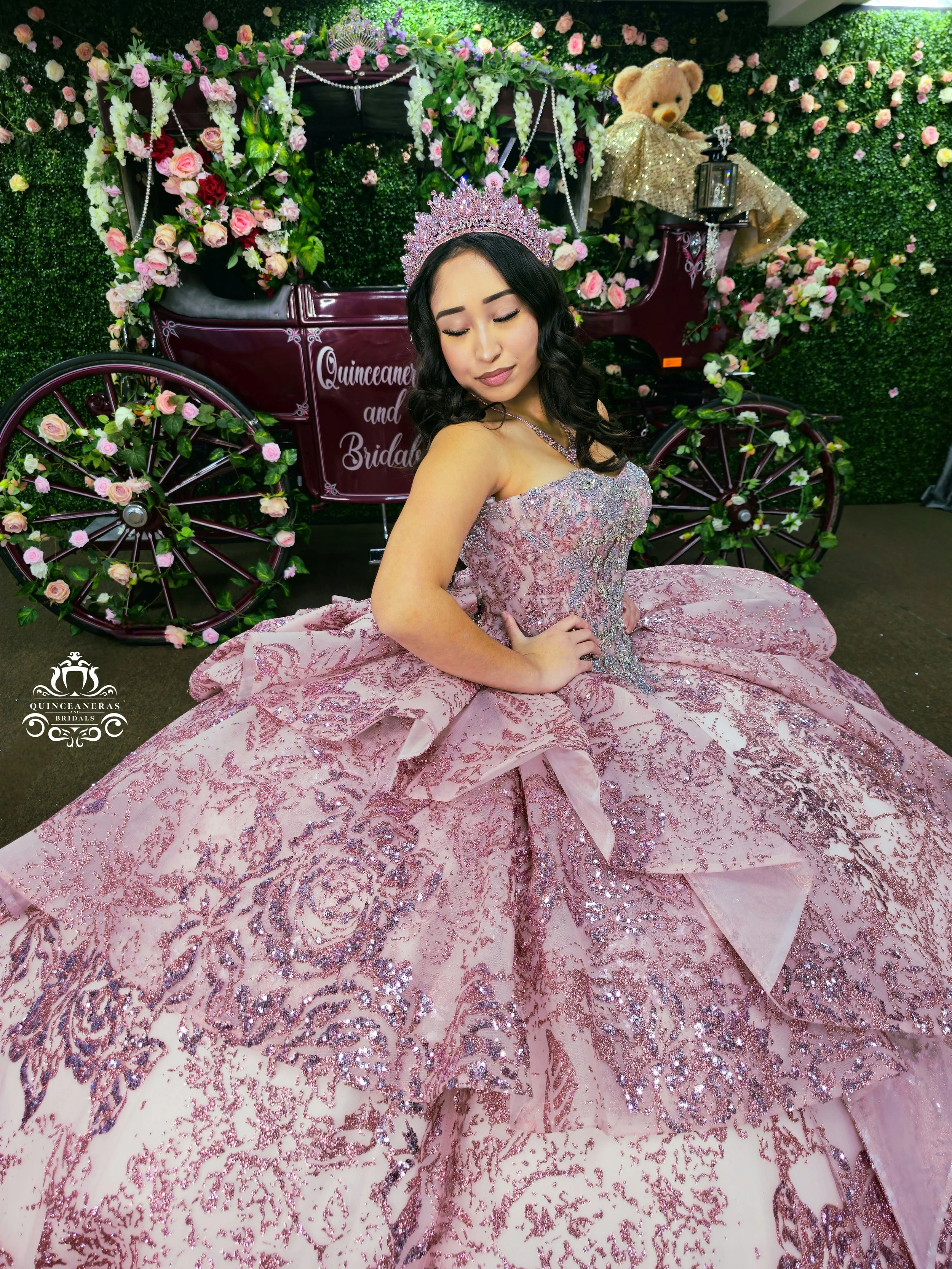 A woman in a pink, sparkly quinceañera dress with intricate embroidery and a large, flowing skirt, standing in front of a decorated carriage with flowers, wearing a tiara and jewelry.