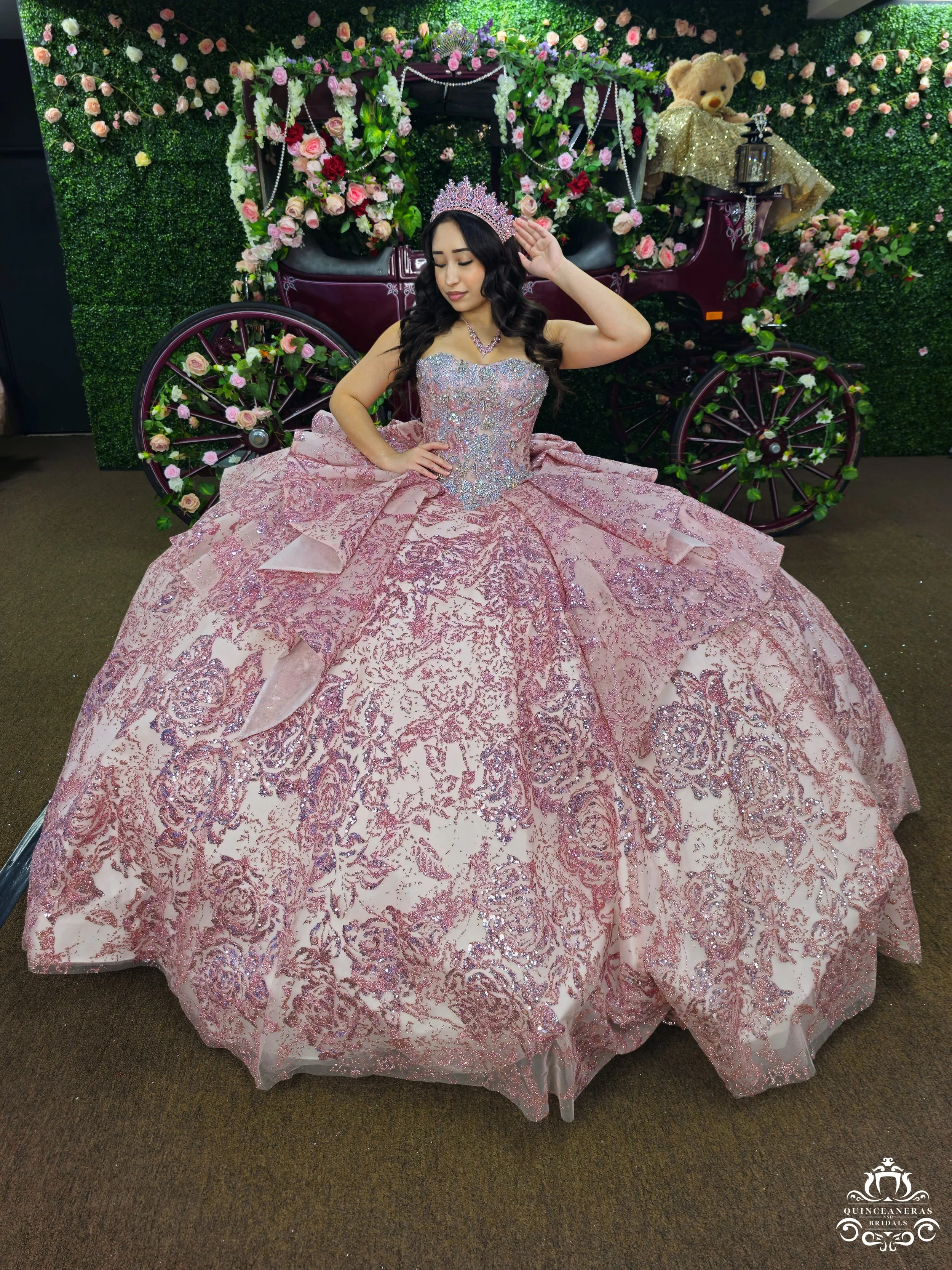 A woman in a pink and silver quinceanera dress with a tiara and necklace, posing in front of a floral carriage with a large teddy bear on top, surrounded by a green floral wall with pink roses.