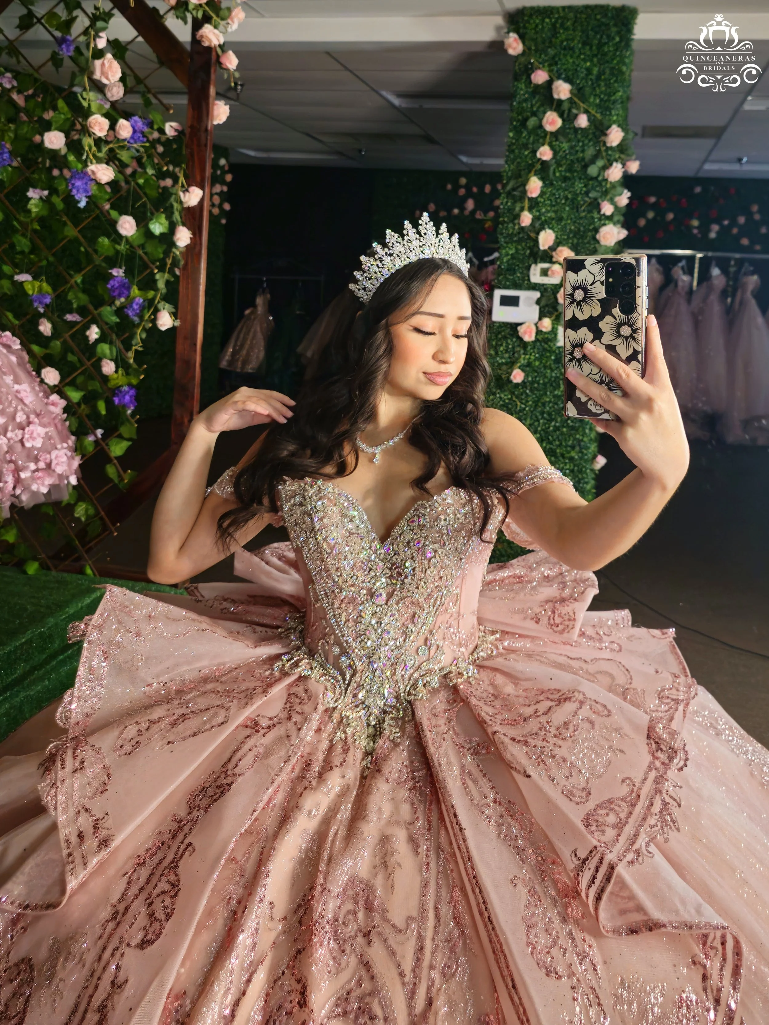 Young woman taking a selfie in a pink, ornate quince gown with a tiara, surrounded by floral decorations.