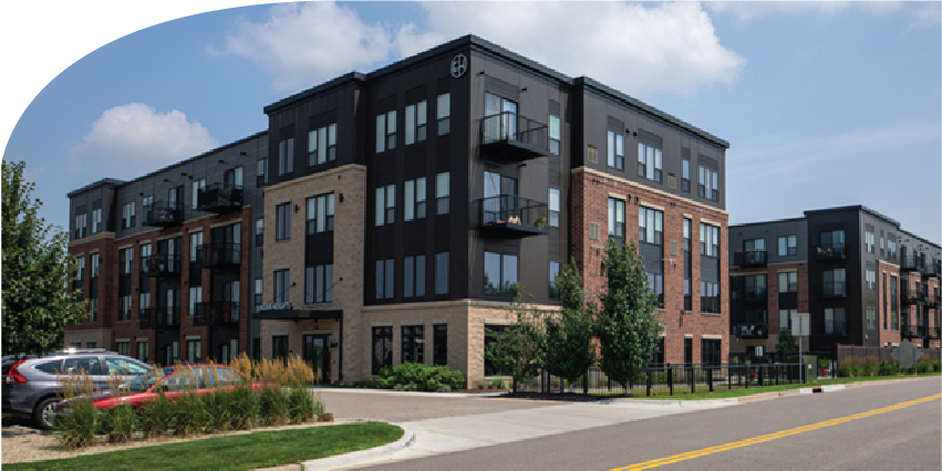 Modern four-story apartment building with brick and dark siding, featuring balconies, in a suburban area with landscaped front yards and parked cars.