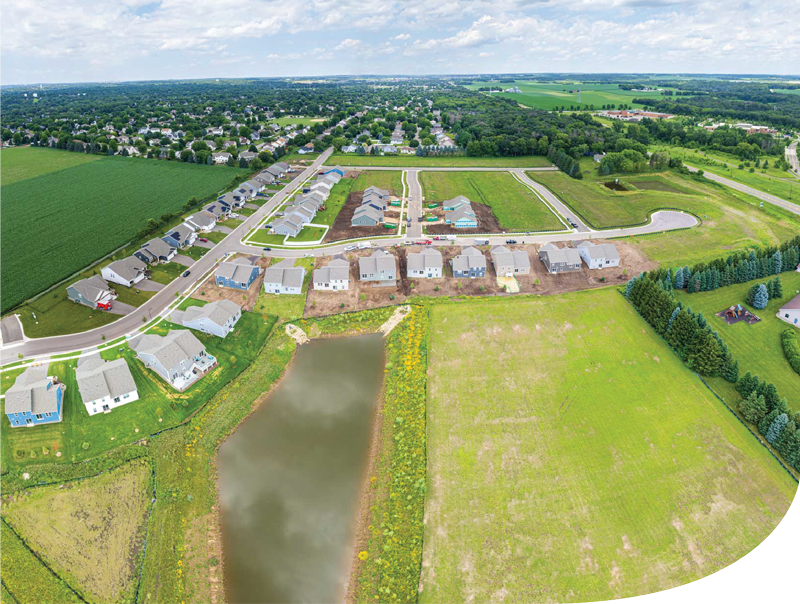 Aerial view of a suburban neighborhood under construction, with houses, roads, a pond, and surrounding green fields.
