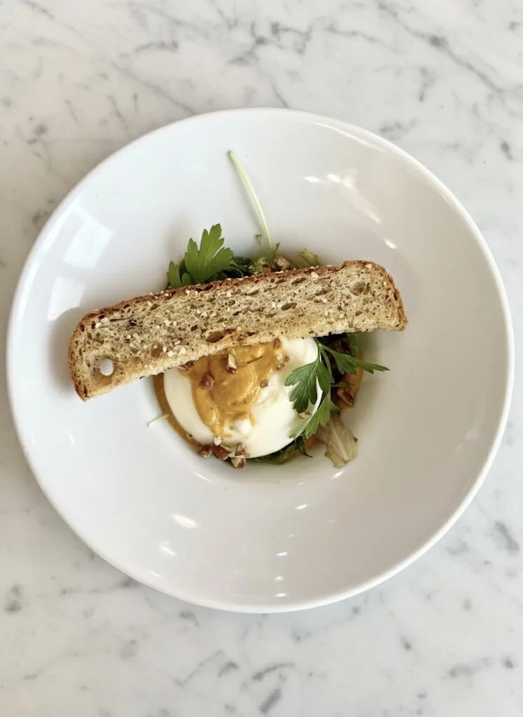 Poached egg topped with sauce and chopped nuts, garnished with cilantro and served with a slice of whole grain bread on a white plate.