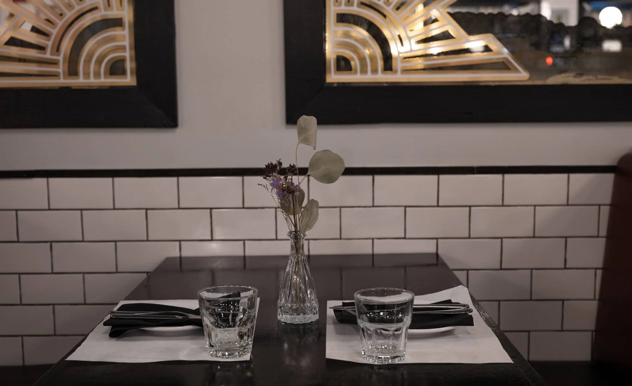 A restaurant table set with two glasses of water, black napkins, and utensils, and a small vase with dried flowers, against a backdrop of white subway tile walls and framed modern artworks.