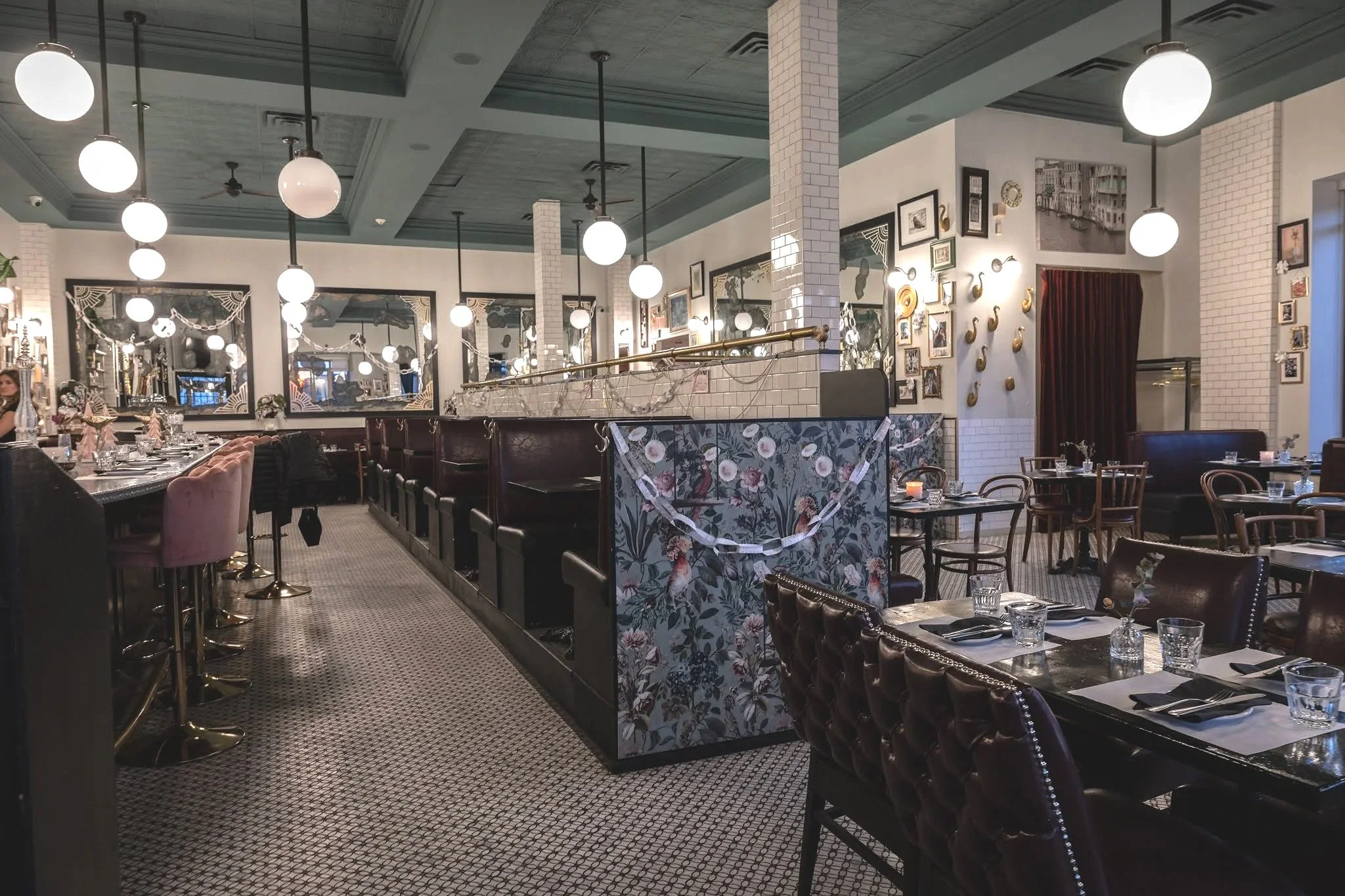 Interior of a stylish restaurant decorated with framed pictures on the walls, pendant lights hanging from the ceiling, and a mix of chairs and booths set up around tables with glassware and cutlery. art deco vibe. modern decor. chic look. events grou
