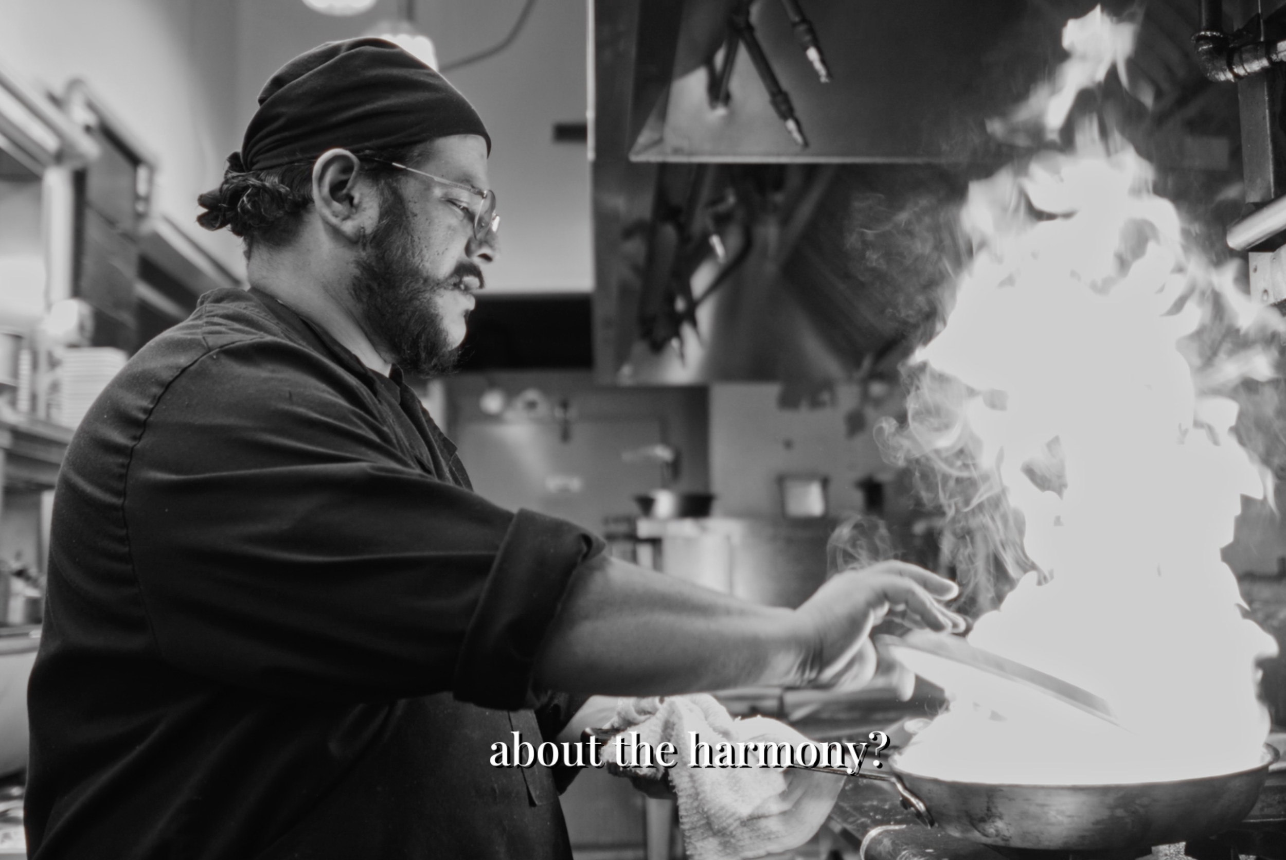A chef cooking in a professional kitchen with a flame coming from a pan, wearing glasses and a chef's hat, with the text 'about the harmony?' overlaid on the image.