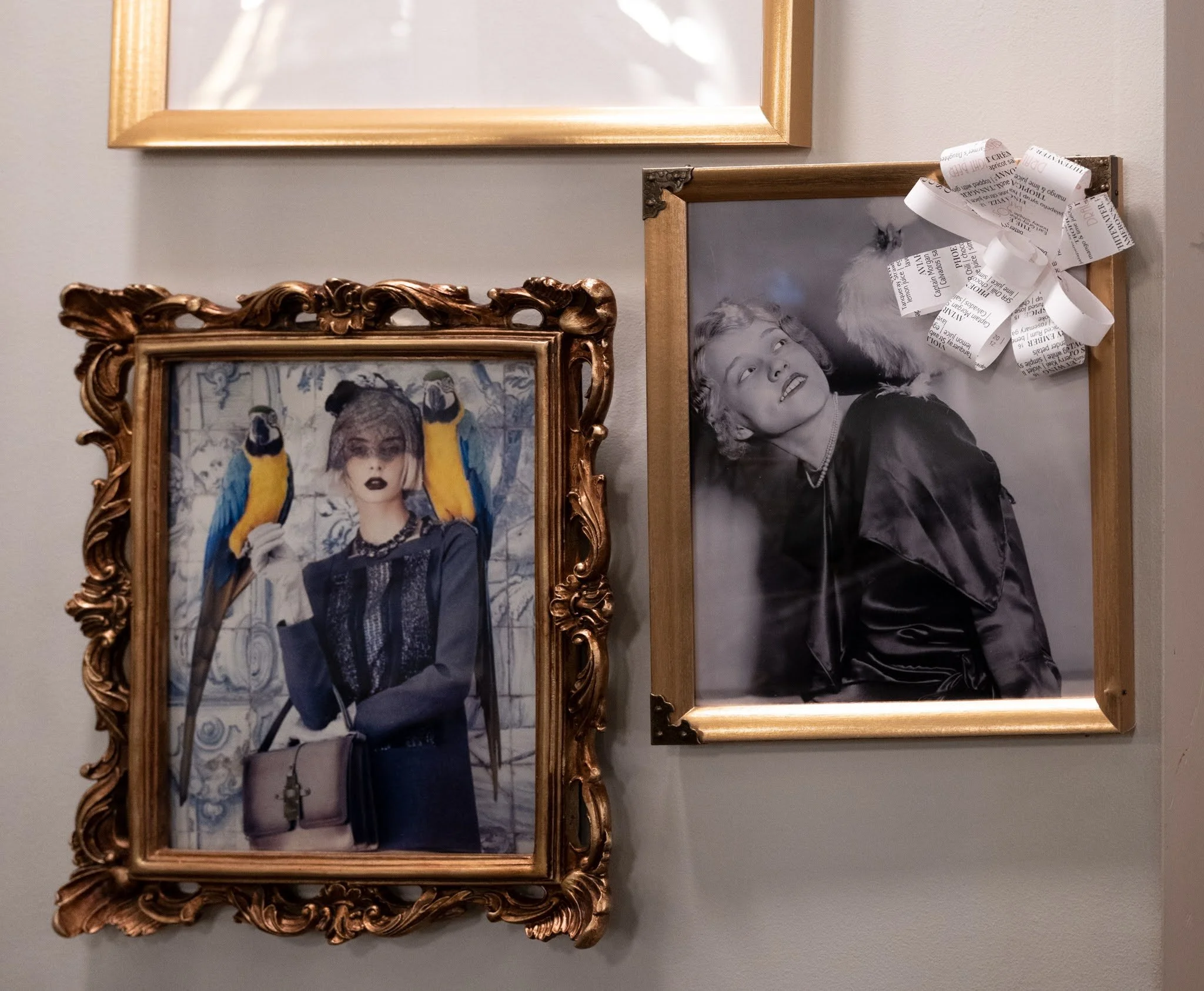 Two framed photographs of women on a wall. The left photo shows a woman with dark lipstick, wearing dark clothing, and two colorful parrots perched on her shoulders. The right photo is a black-and-white portrait of a woman looking up, with a fluffy d