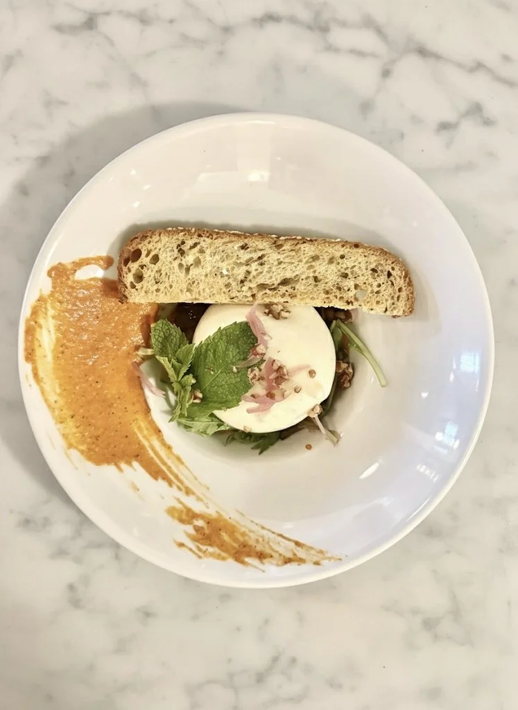 Poached egg with greens and pink pepper flakes, with a slice of bread and a swirl of orange sauce on a white plate.