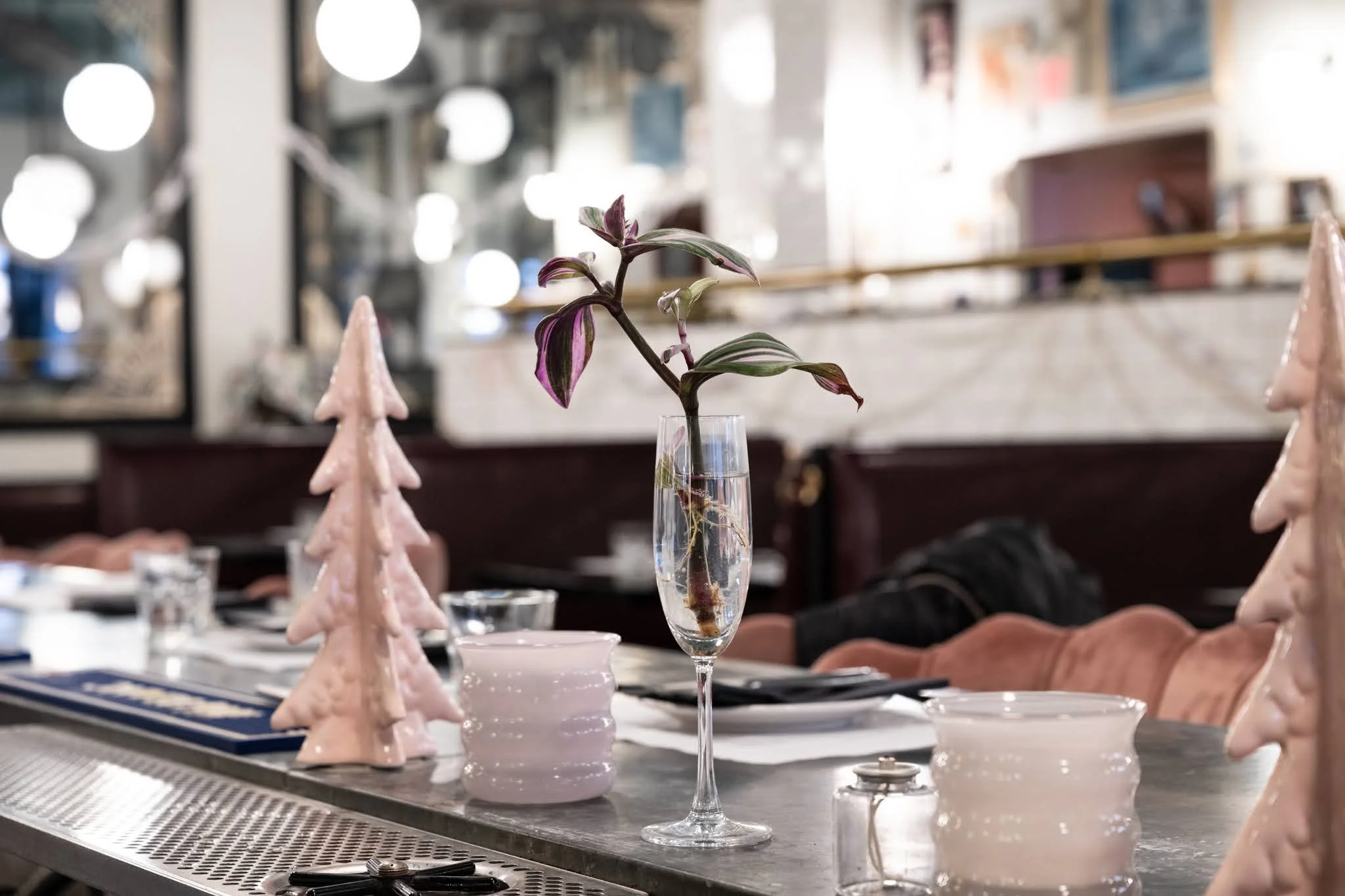 A dinner table decorated with a tall glass vase with a plant, pink ceramic Christmas trees, white ceramic containers, black napkins, and utensils, in a restaurant setting with blurred background. The atmosphere is lively, with soft jazz music playing