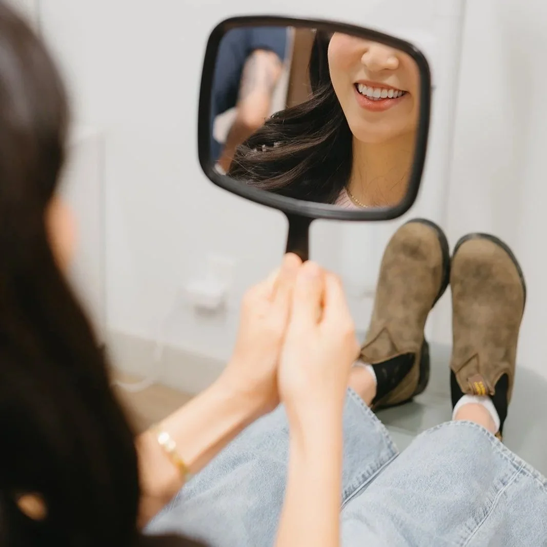 Person sitting on the floor looking into a hand mirror, smiling at their reflection, wearing jeans and brown boots.