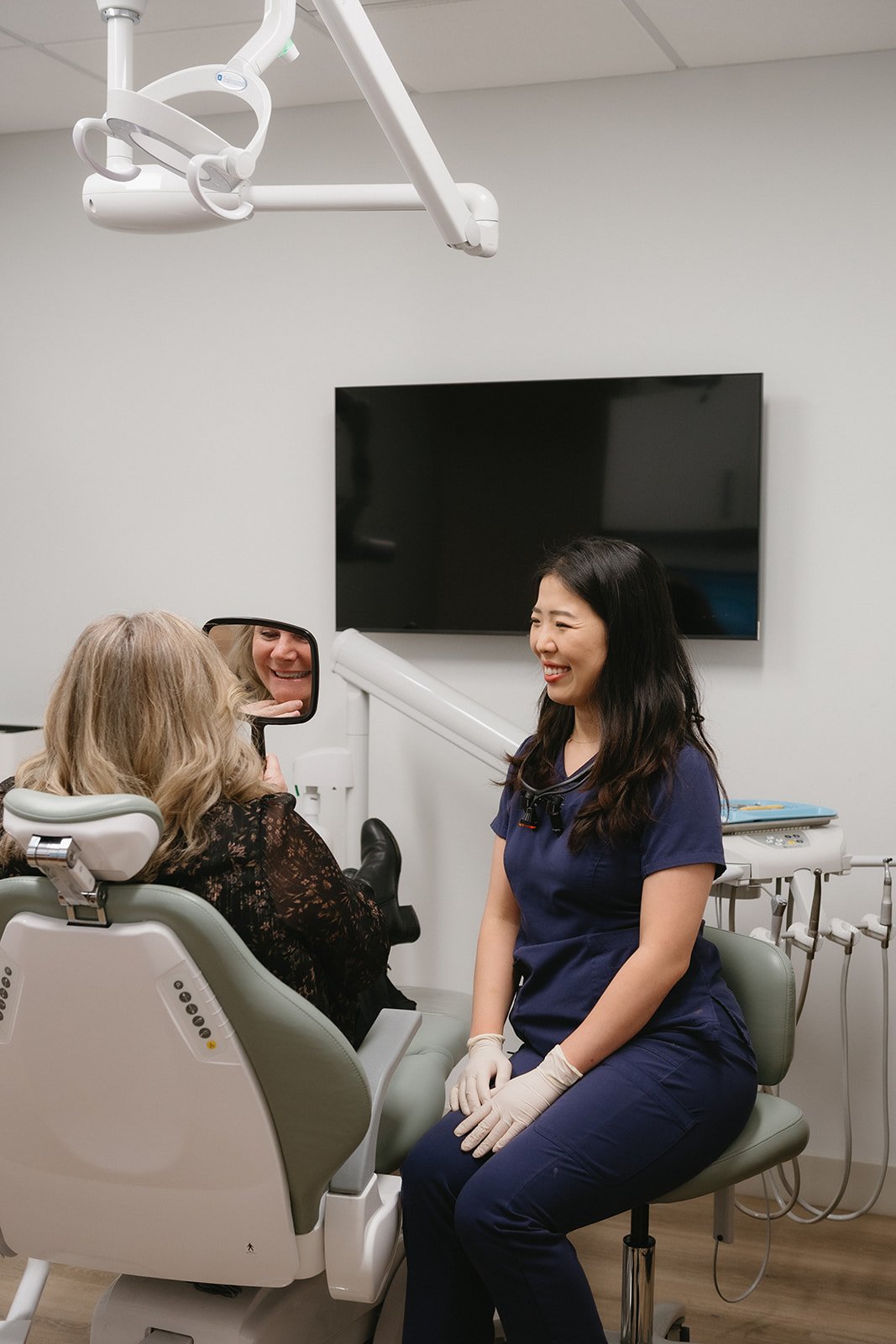 A female dental professional in navy scrubs and gloves smiling at a patient in a dental chair, with a mirror reflecting the patient's face. The setting is a modern dental clinic with medical equipment and a large monitor on the wall.