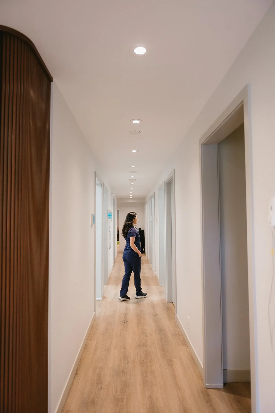 A nurse standing in a hallway of a medical facility, wearing dark blue scrubs and looking to the side.