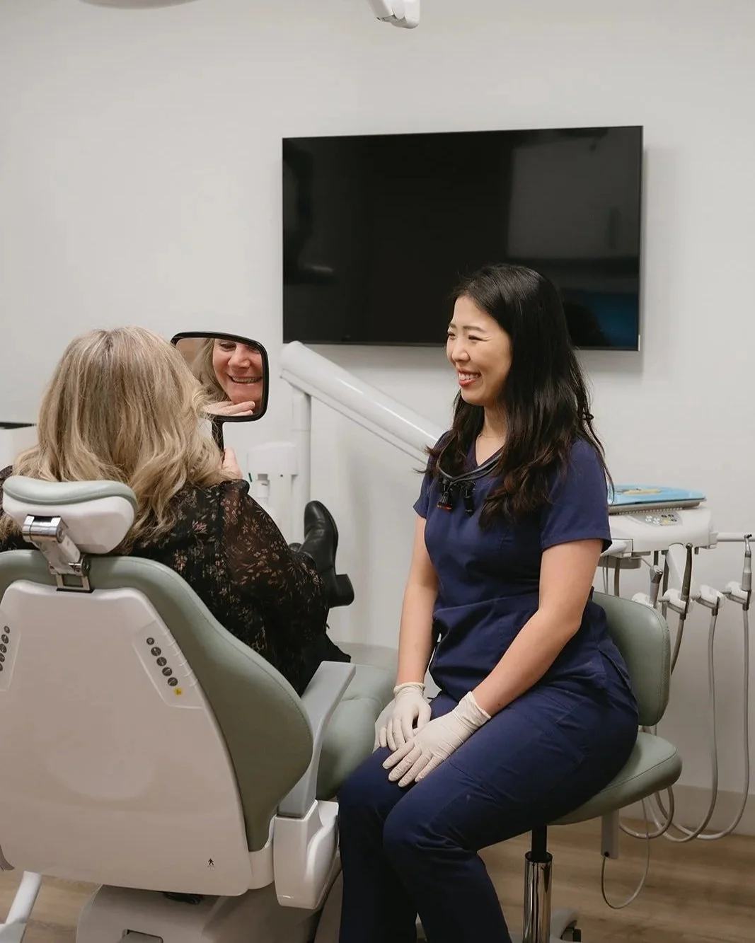 A dentist smiling and talking with a patient in a dental office, with both women smiling. The patient is sitting in a dental chair and looking into a mirror.