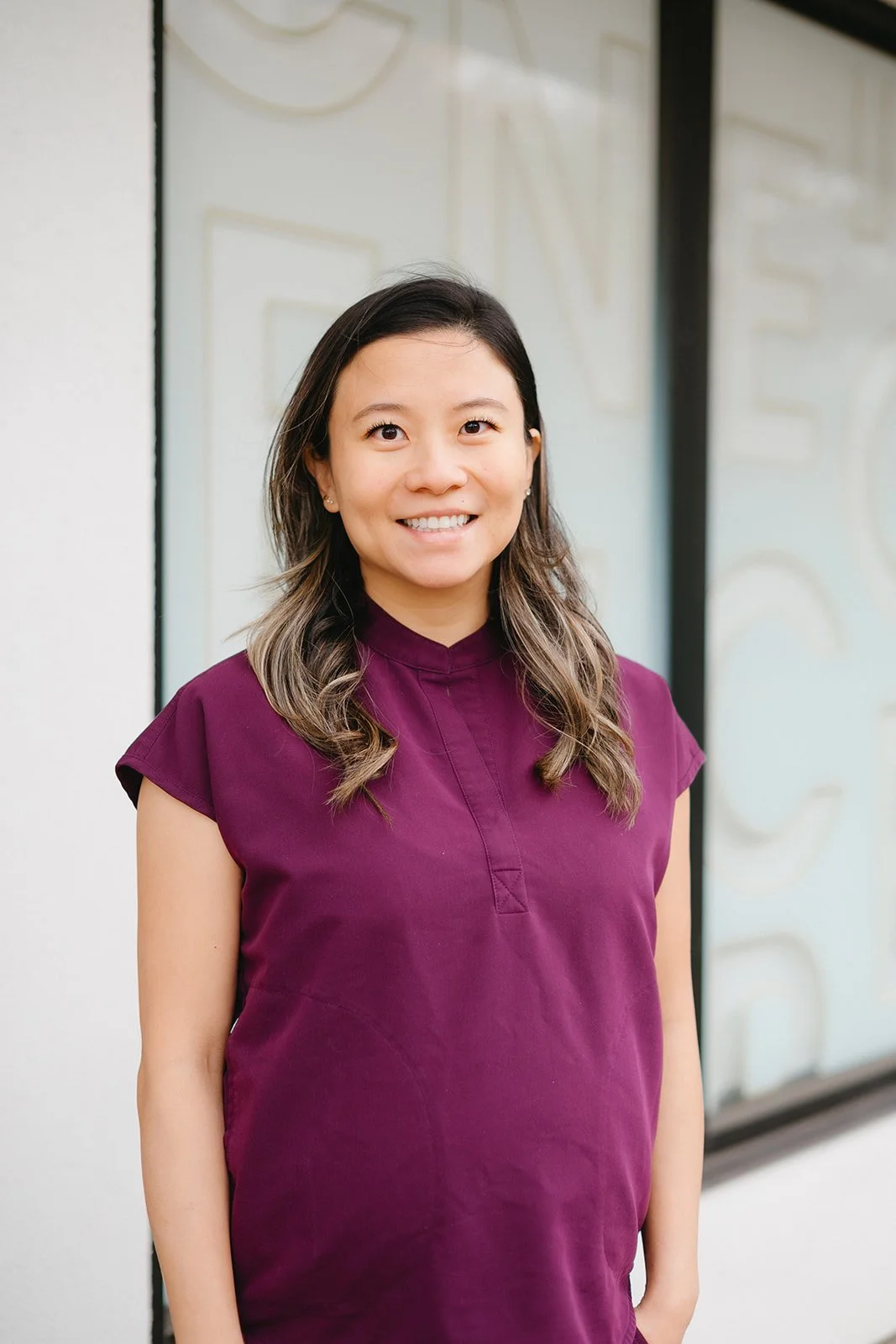 A woman with shoulder-length wavy hair wearing a purple top, smiling, standing outdoors in front of a large glass wall with partial blurred text.