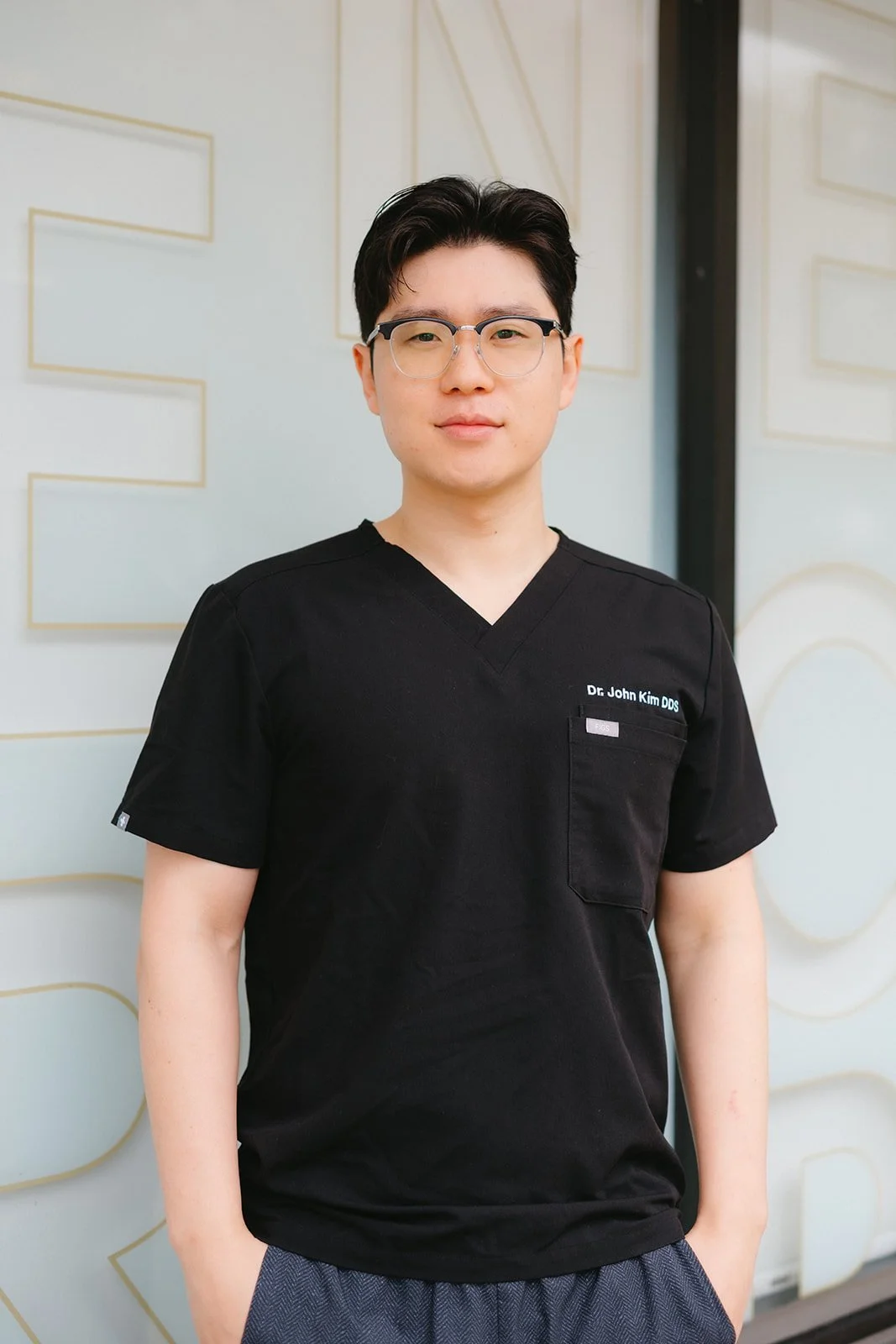 A young man wearing glasses and a black medical scrub top standing in front of a light-colored wall with decorative gold lines.