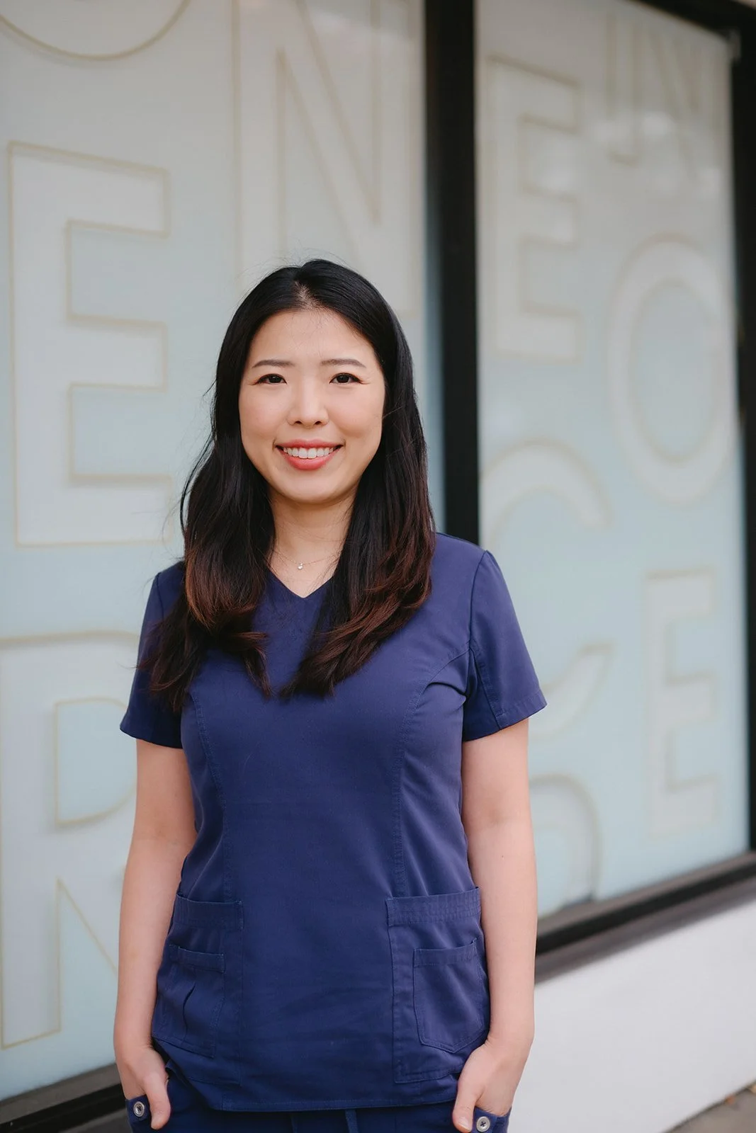 A woman wearing navy scrubs standing outside in front of a light-colored wall with large raised letters.