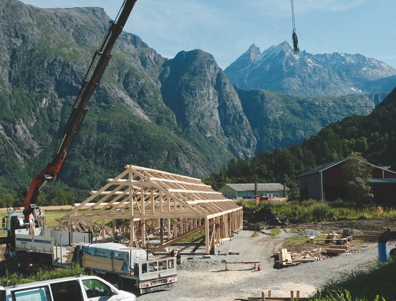 Construction site in a mountainous area with a wooden framework of a building, a crane, and construction vehicles, surrounded by green trees and rugged mountains in the background.