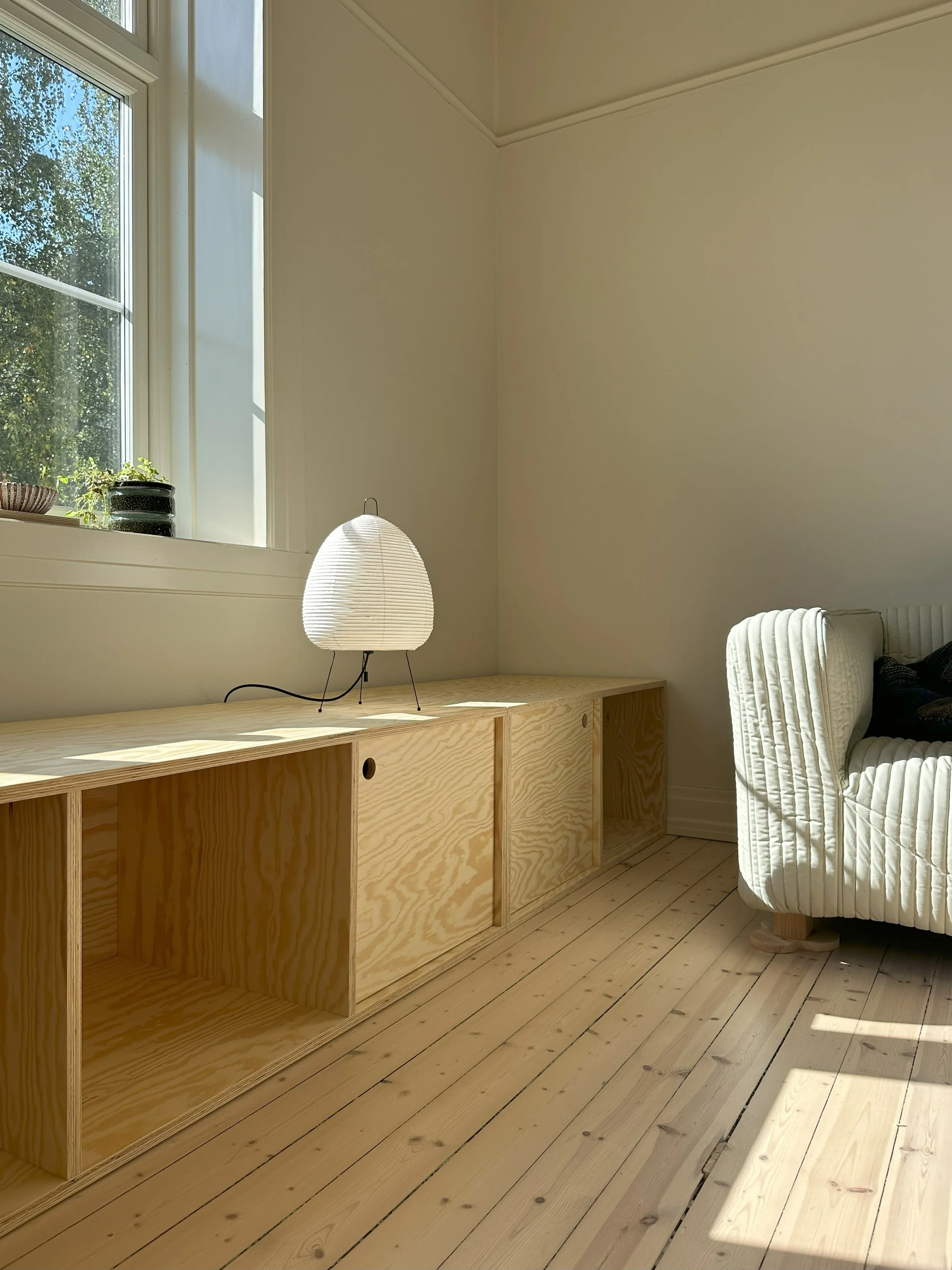 A minimalist living room corner with a wooden cabinet, a white paper lantern lamp, a window with sunlight and plants outside, and a white upholstered sofa.