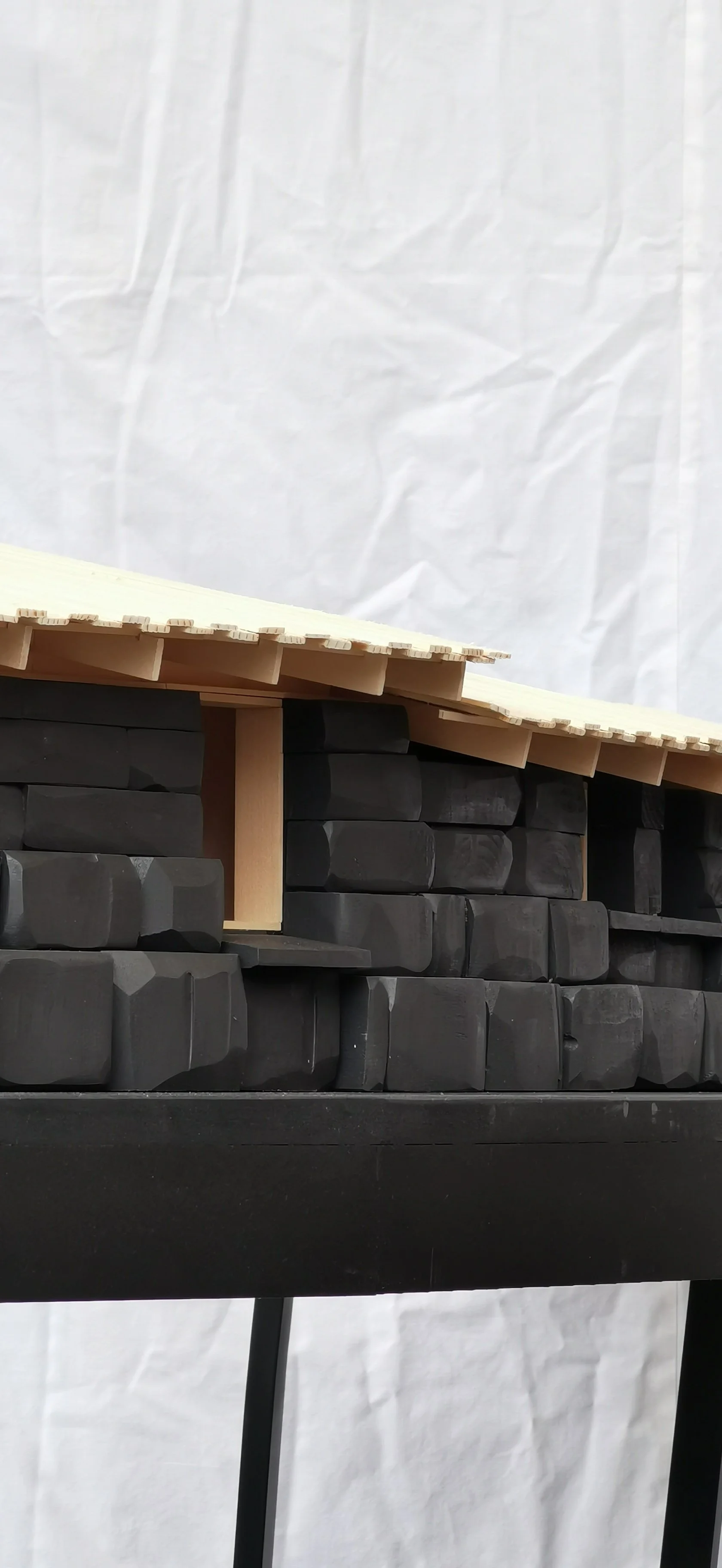 A miniature architectural model with black brick-like structures, a small archway, and a sloped beige roof, placed on a black stand.