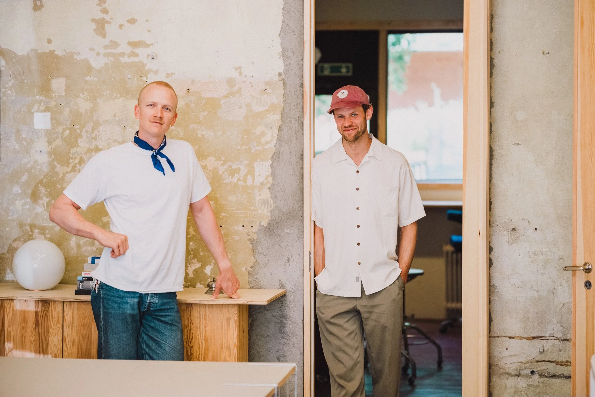 Two men standing in a room with a distressed wall; one is leaning on a wooden surface with mental items and the other is standing in a doorway wearing a hat and casual shirt.