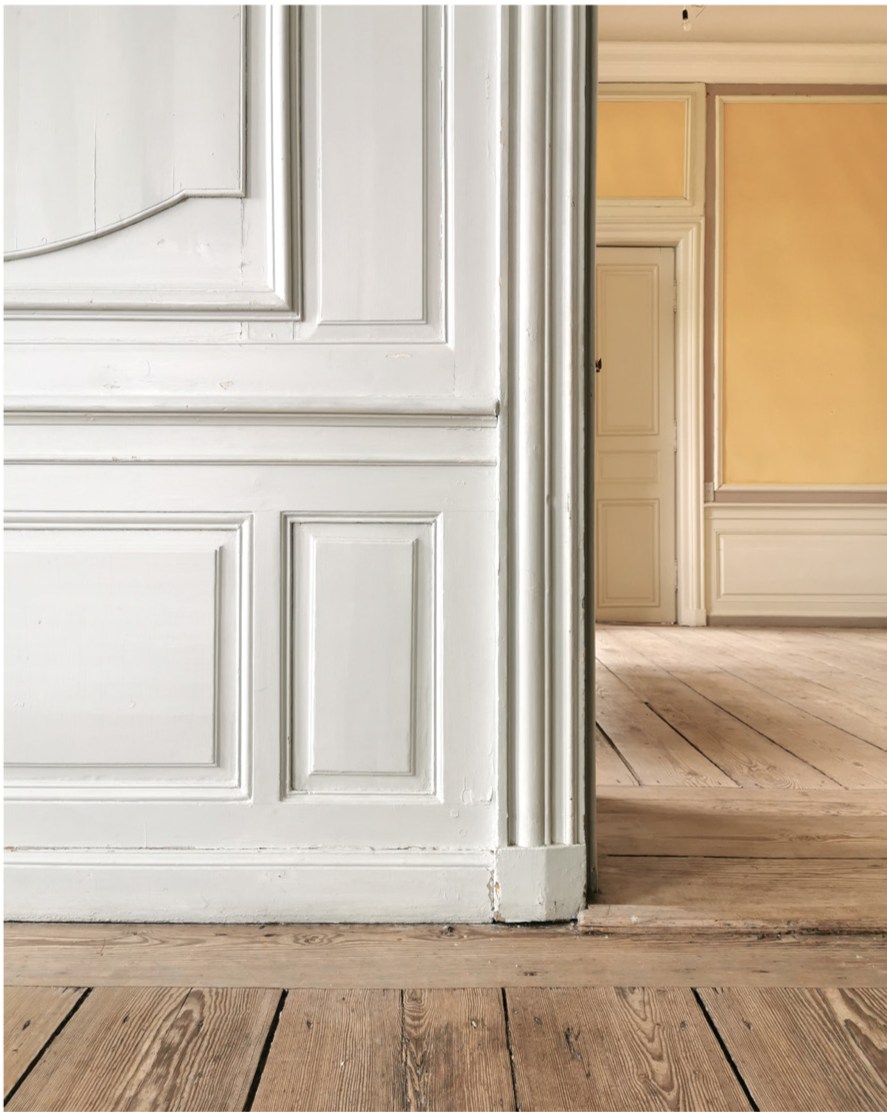 Close-up of a white wooden door frame with detailed molding, partially open to reveal a room with yellowish walls and a wooden floor in the background.