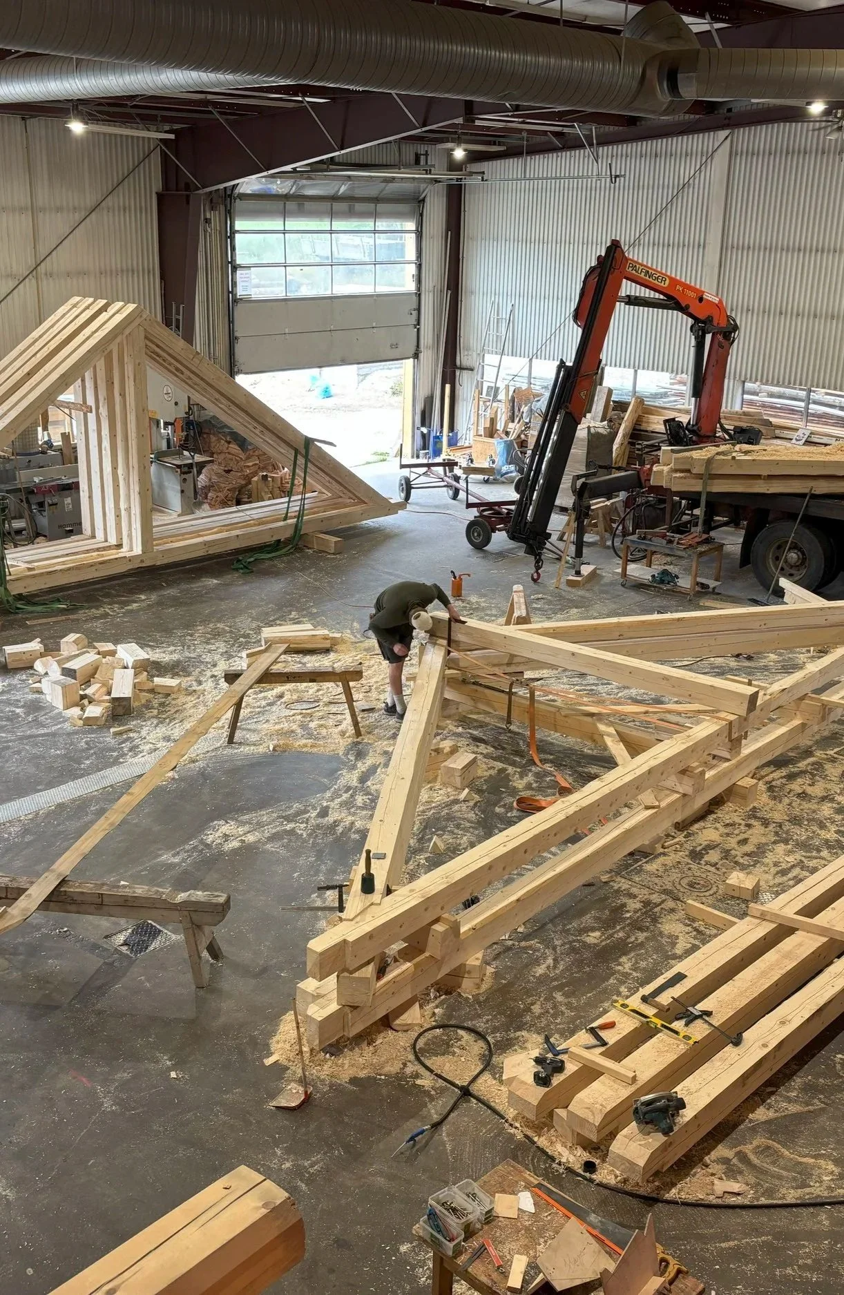 Interior of a woodworking workshop with a partially built triangular wooden structure, a worker measuring wood, and various construction tools and materials scattered around.