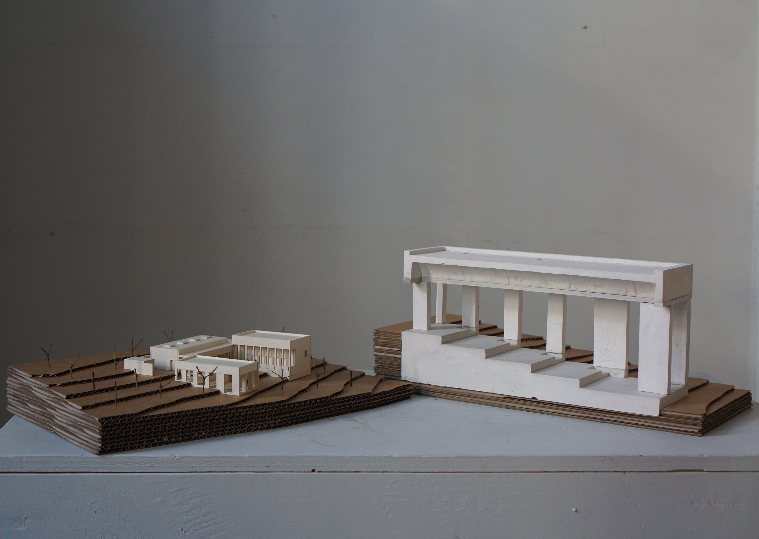 Architectural models of modern buildings on layered cardboard bases, one on the left and the other on the right, against a plain gray background.