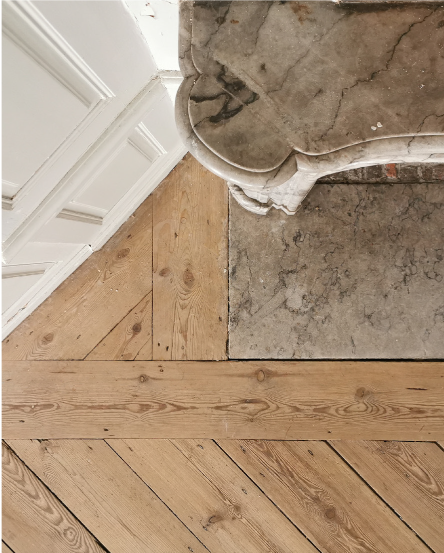 A close-up of a room's corner showing different flooring types: white painted wood paneling on the wall, a narrow strip of wood flooring, a section of uneven concrete, a vintage marble fireplace, and honey-colored wood flooring.