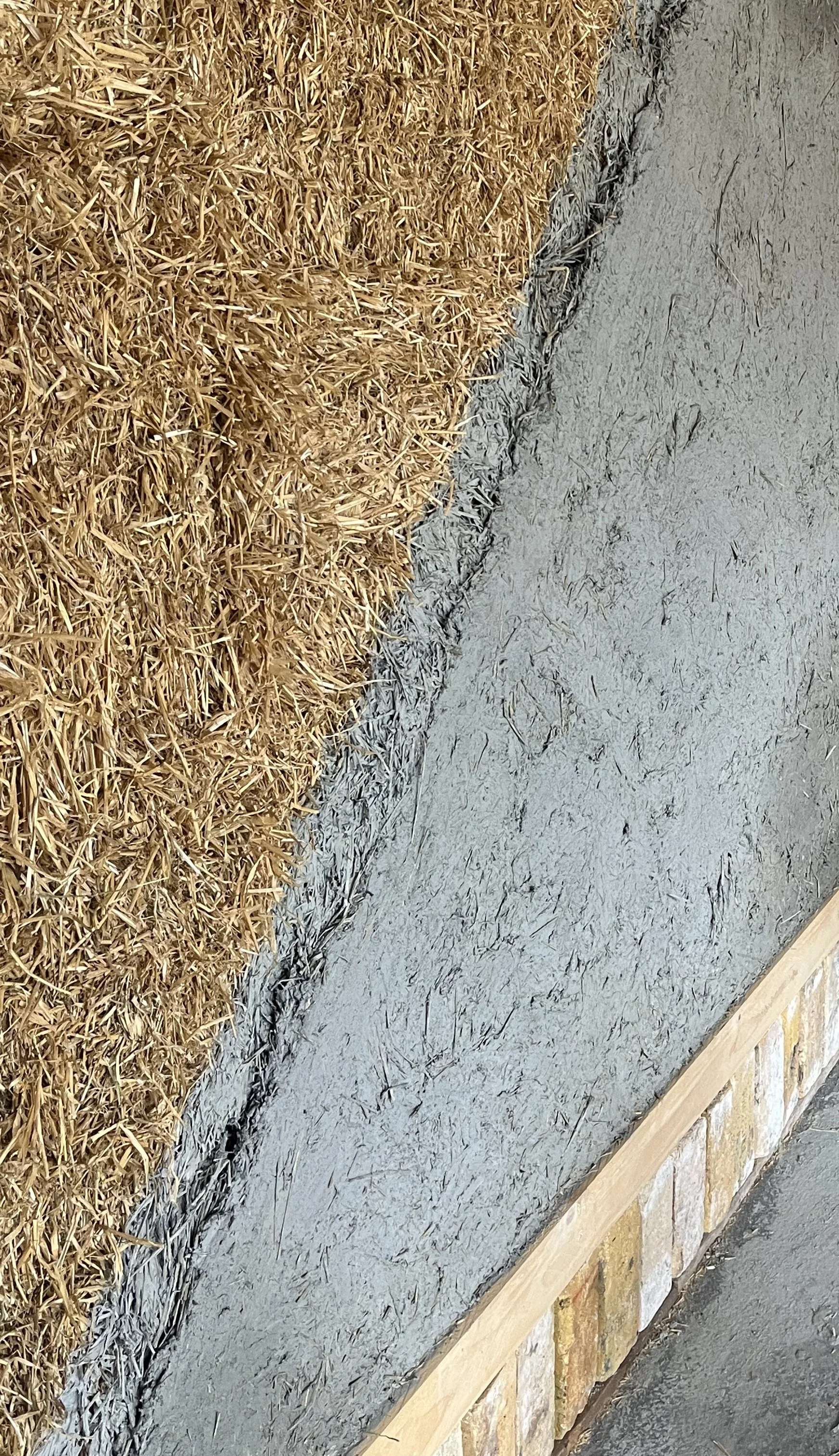 Close-up view of a concrete wall with freshly applied wet cement, next to a soil and straw-covered area, with a wooden border at the bottom.