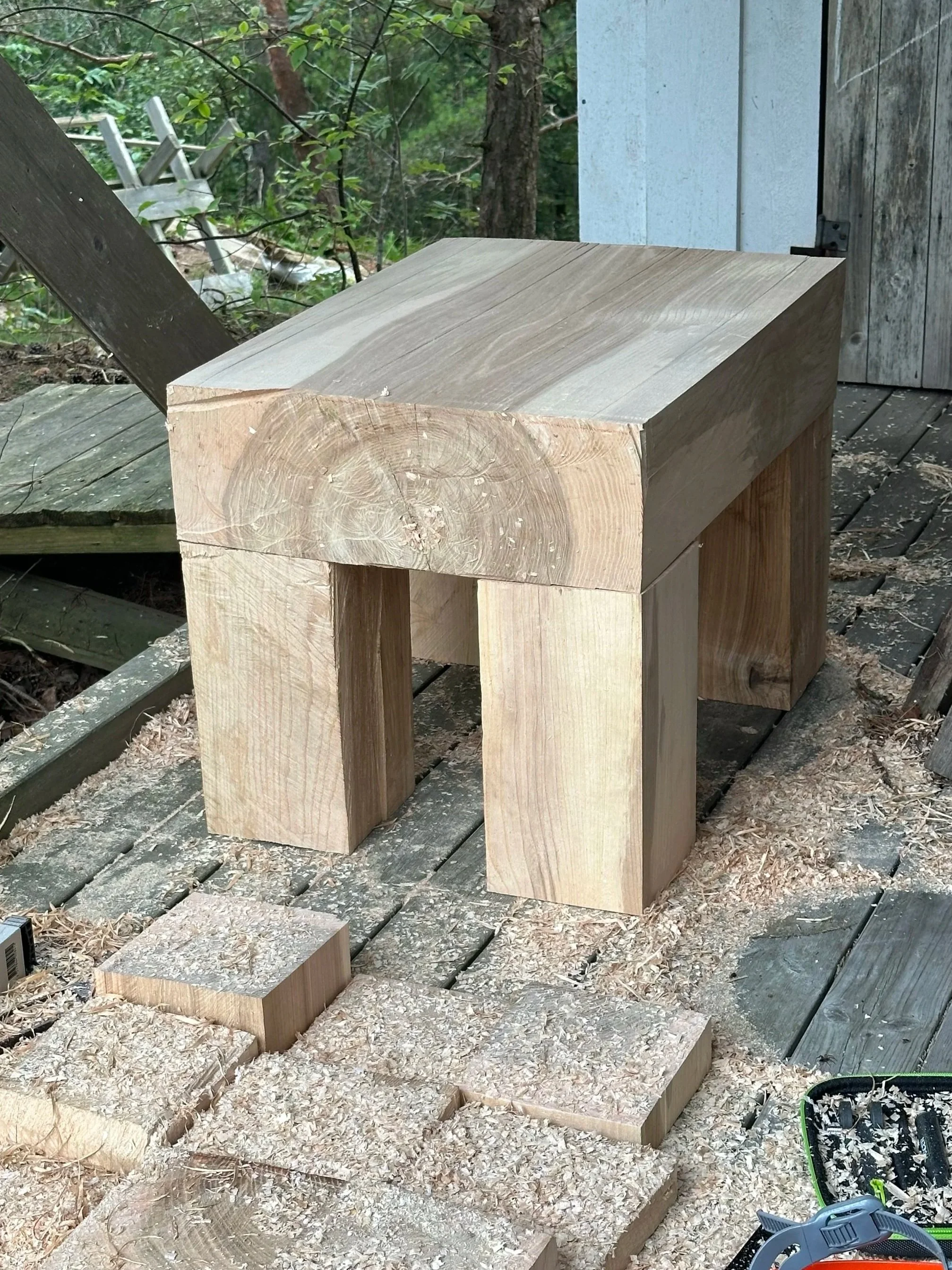 A partially assembled wooden stool outdoors on a wooden deck with wood shavings and pieces around, surrounded by trees and forest.