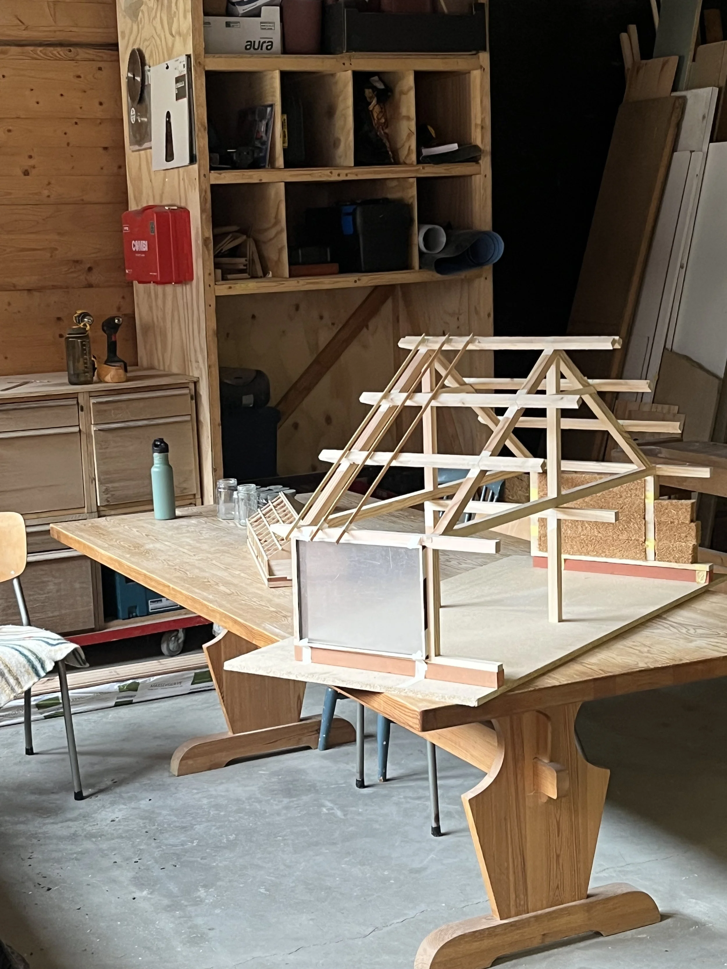 A woodworking workshop showcasing a table with a detailed wooden architectural model or miniature structure on top, shelves with various tools and materials in the background, and a chair with a striped cloth in the foreground.