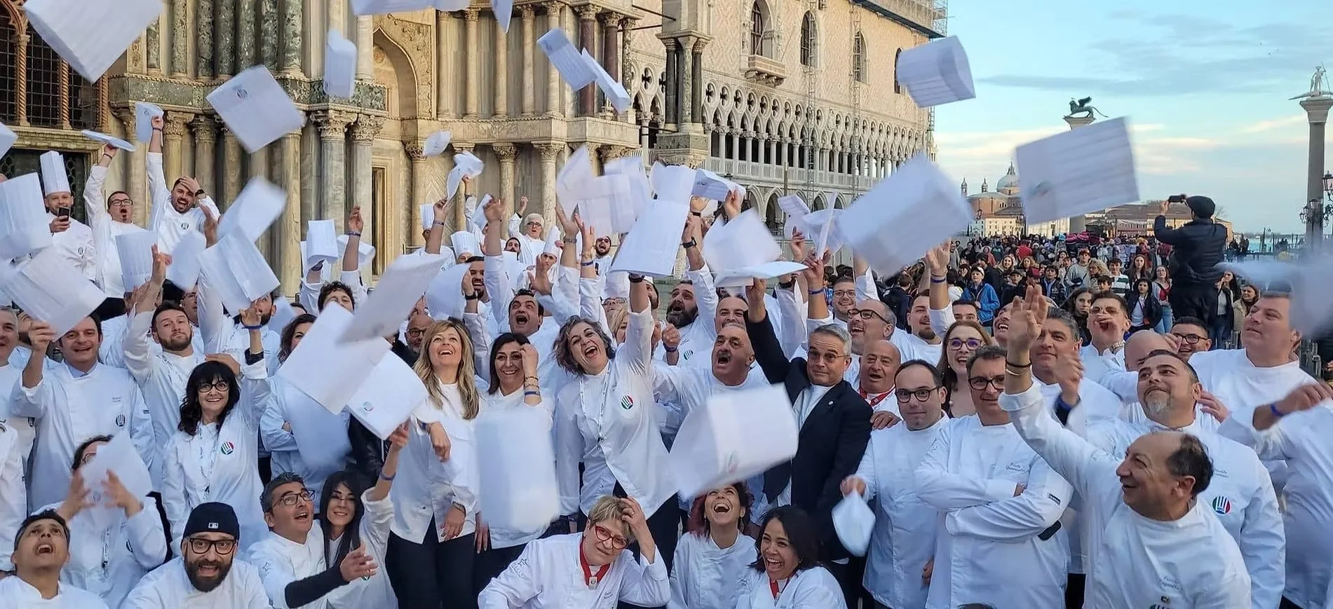 Group of chefs and culinary students celebrating and throwing papers in the air outside, with historic buildings in the background.