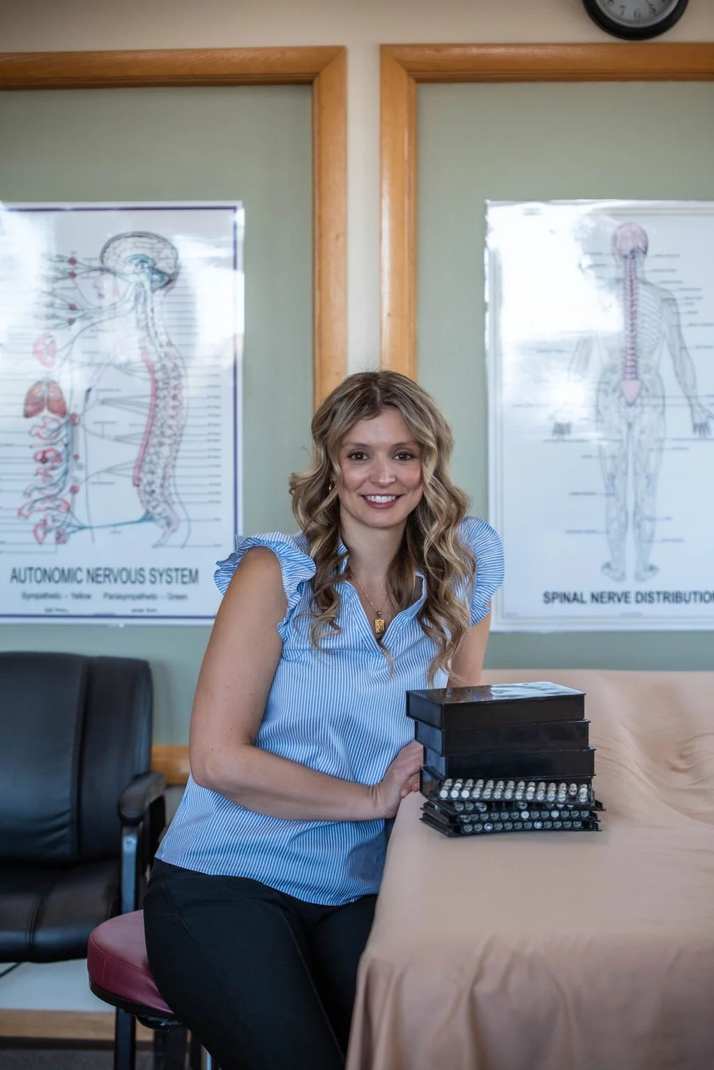 A smiling woman with blonde wavy hair and a blue striped top sitting at a table with a black box of markers in front of her. Behind her, there are two posters of the human nervous system on a green wall, and a clock is partially visible in the top right corner.