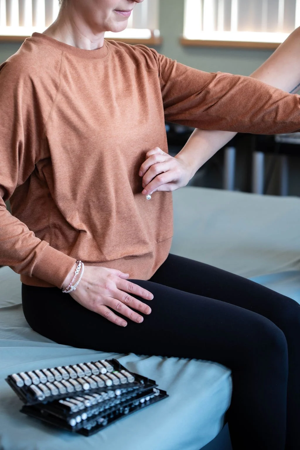 A woman wearing a rust-colored sweatshirt and black leggings sitting on a bed with an assortment of nail polish bottles in front of her, receiving a manicure.
