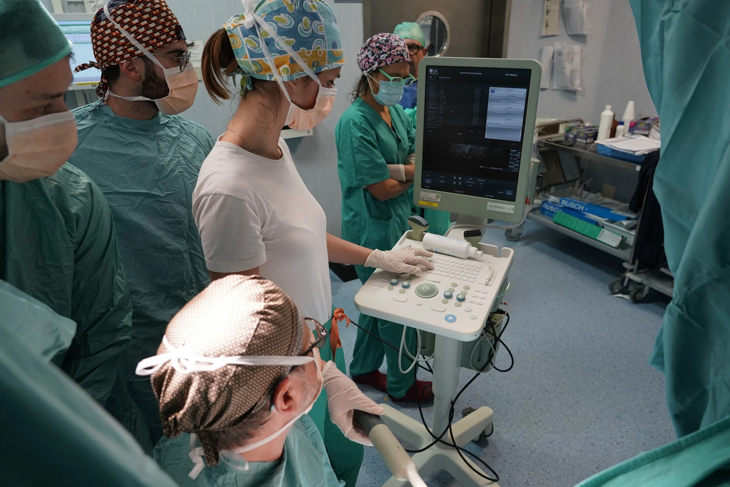 Un gruppo di medici e infermieri in sala operatoria, alcuni indossano maschere e cappelli chirurgici, mentre una donna giovane con cappello colorato e guanti sta usando un monitor di ecografia, con altri che osservano attentamente.