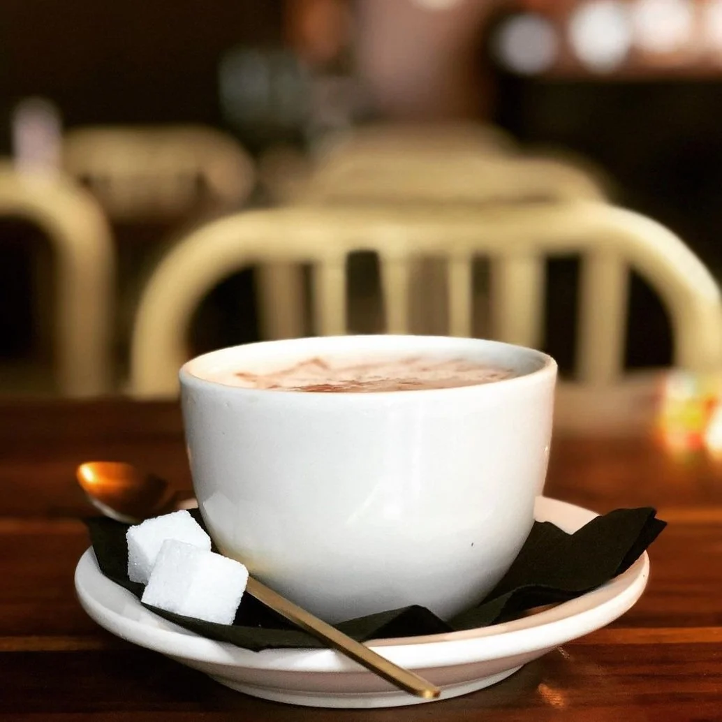 A white cup of hot chocolate topped with whipped cream on a saucer with sugar cubes and a teaspoon, sitting on a wooden table in a cozy cafe.