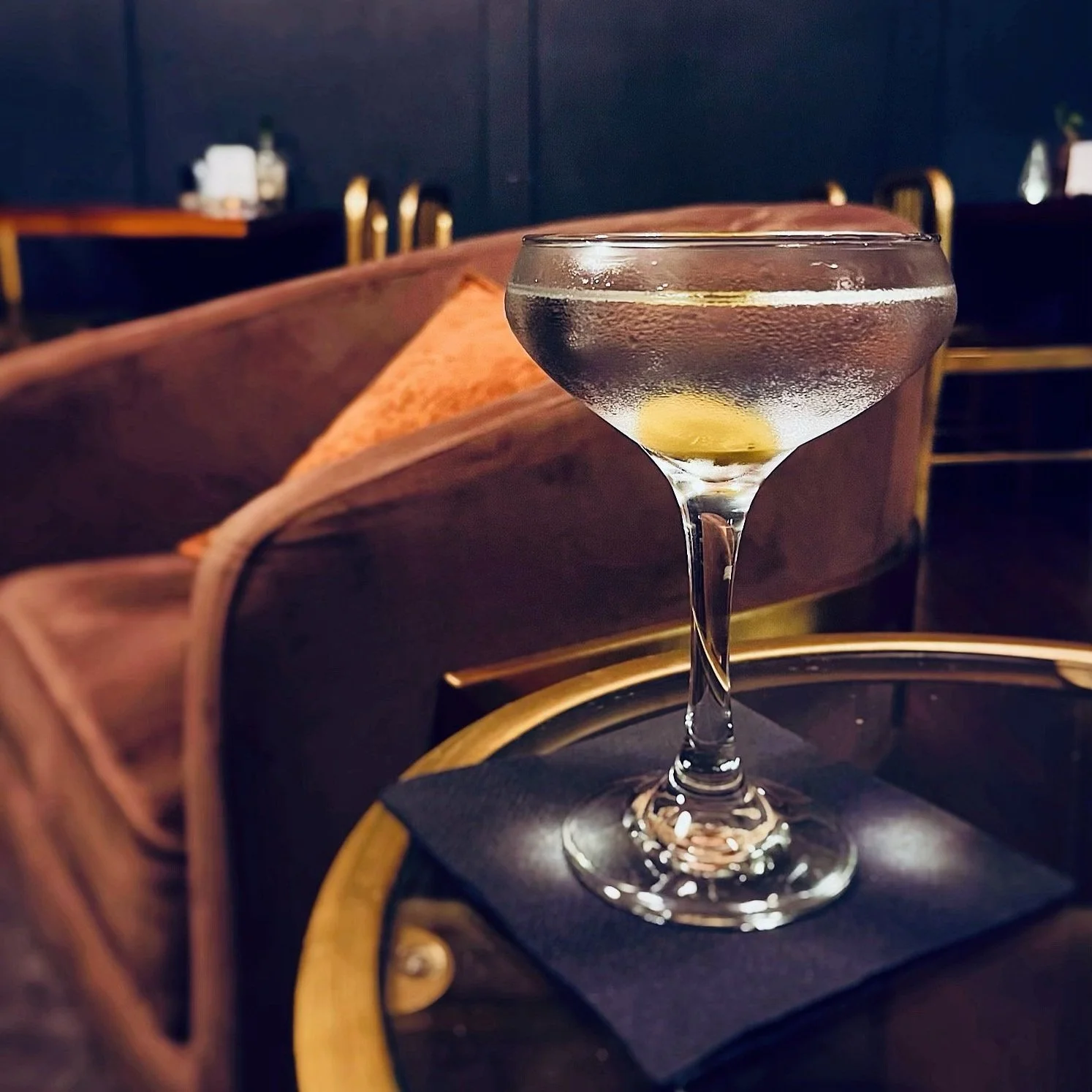 A cocktail glass with a lemon twist on a black napkin on a table in a dimly lit lounge or bar setting.