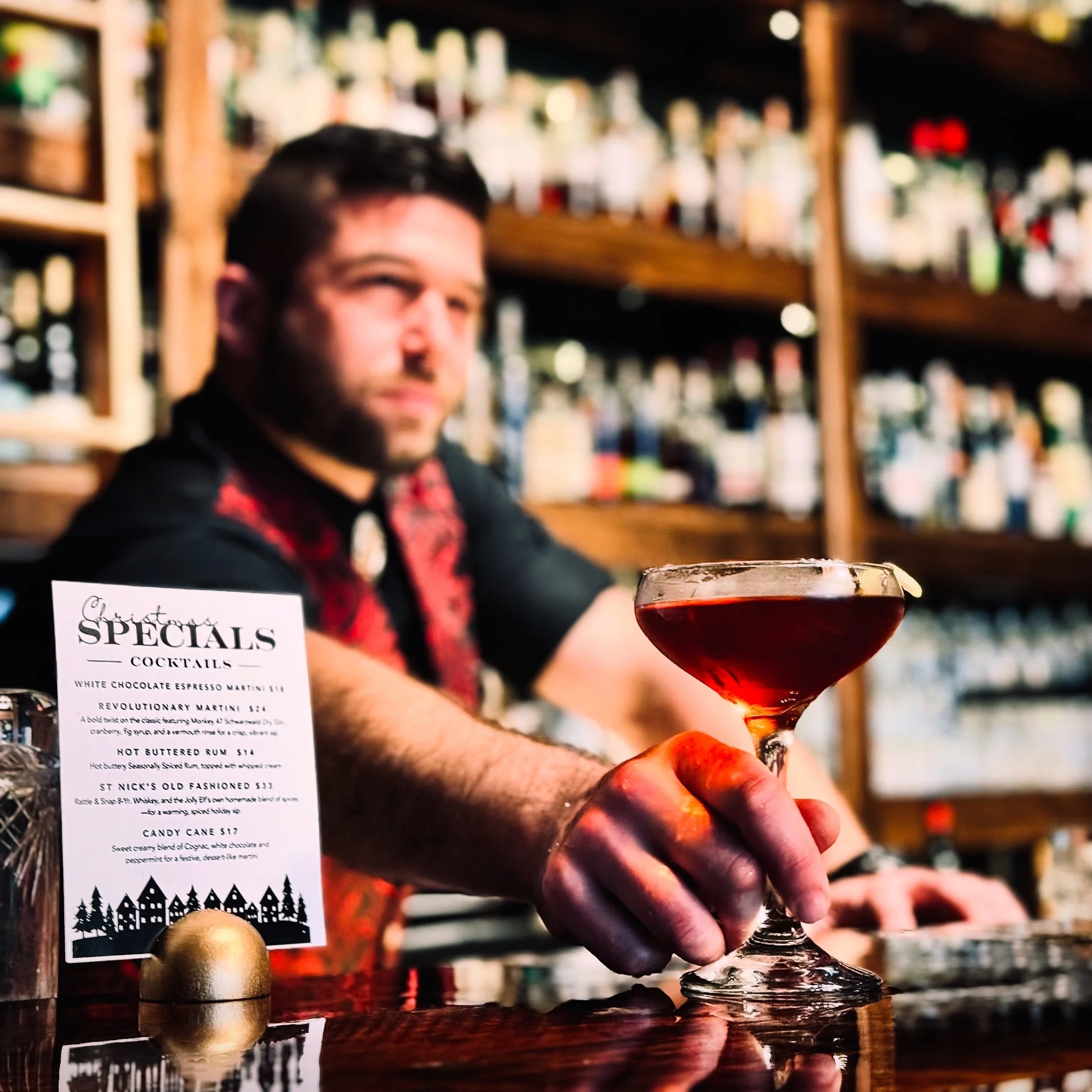 A man behind a bar holding a red cocktail glass in front of a menu and a gold ornament, with shelves of bottles in the background.