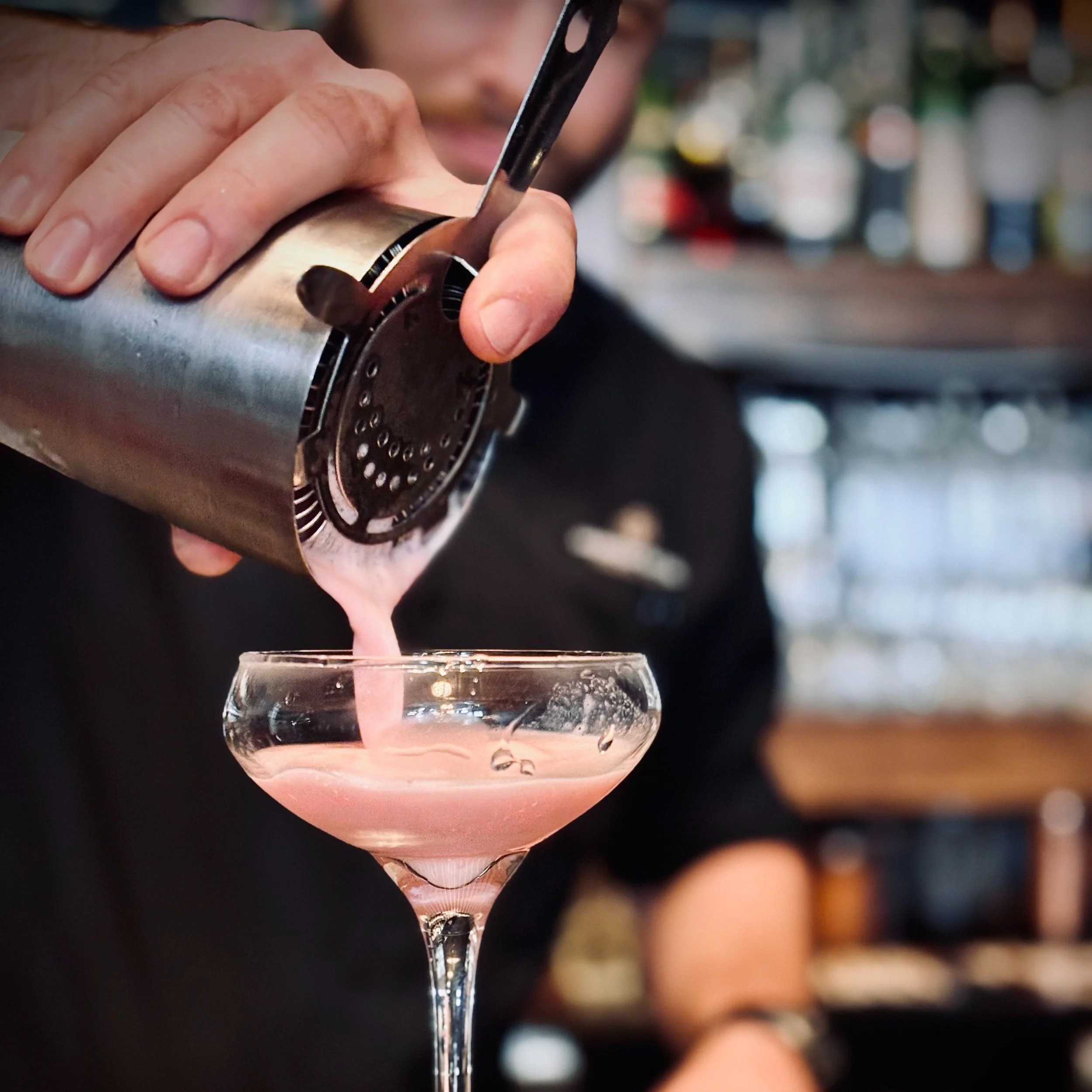 A bartender pours a pink cocktail from a cocktail shaker into a coupe glass, with a blurred bar background.