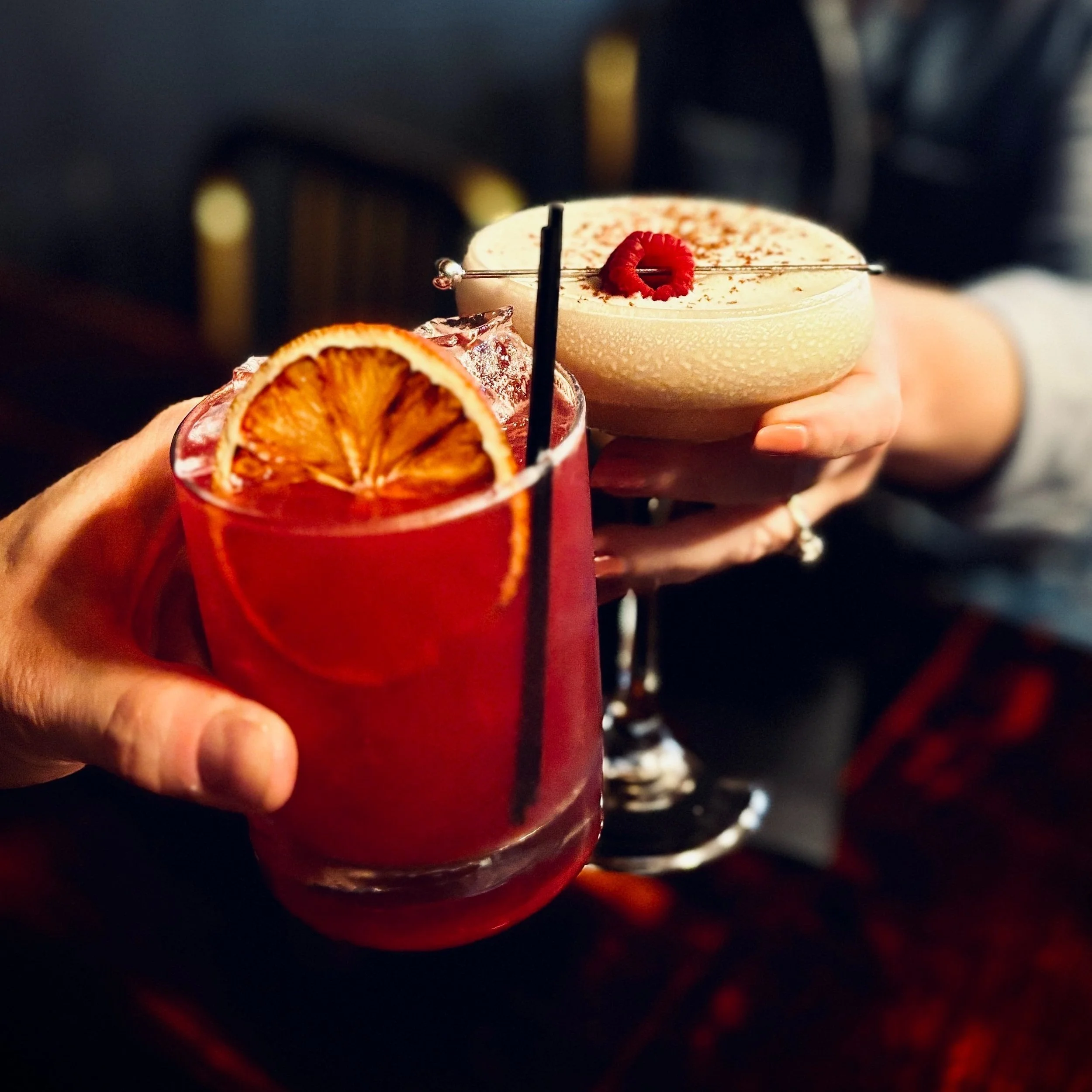 Hands holding two cocktails, one red with dried orange slice and the other creamy with a raspberry garnish, against a blurred dark background.