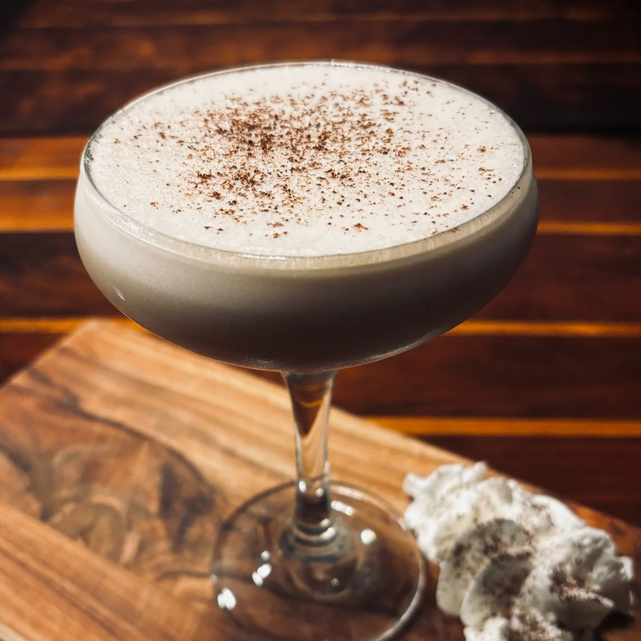 A cocktail in a coupe glass with a frothy top dusted with cocoa powder, placed on a wooden surface next to whipped cream topped with chocolate shavings.