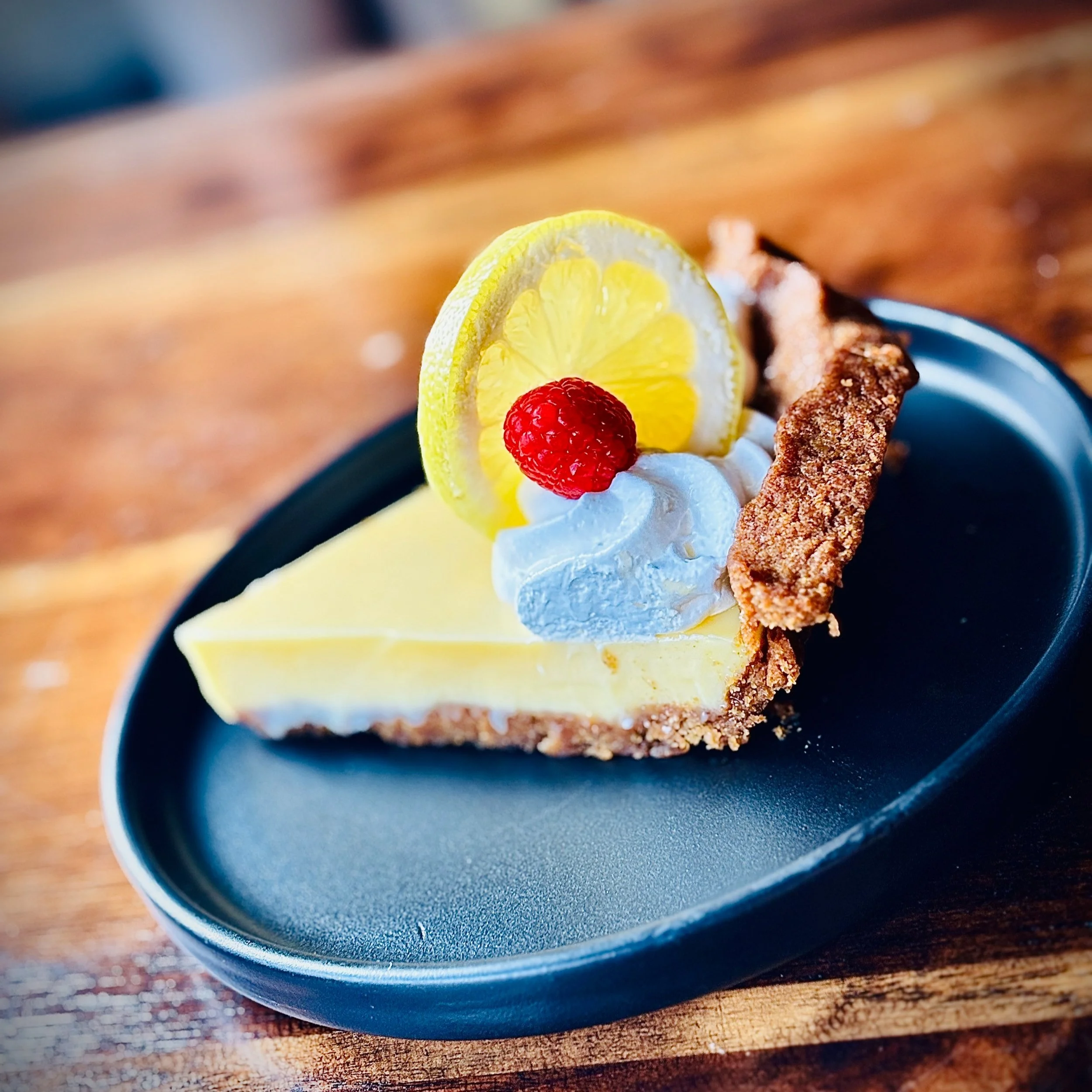 Slice of lemon cheesecake garnished with a raspberry, lemon slice, whipped cream, and a cookie on a black plate.