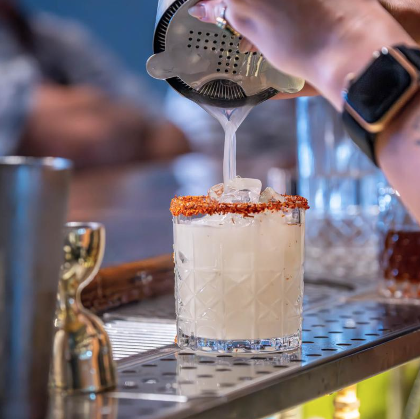 A bartender pouring liquid into a glass with ice, rimmed with chili powder, on a bar counter.