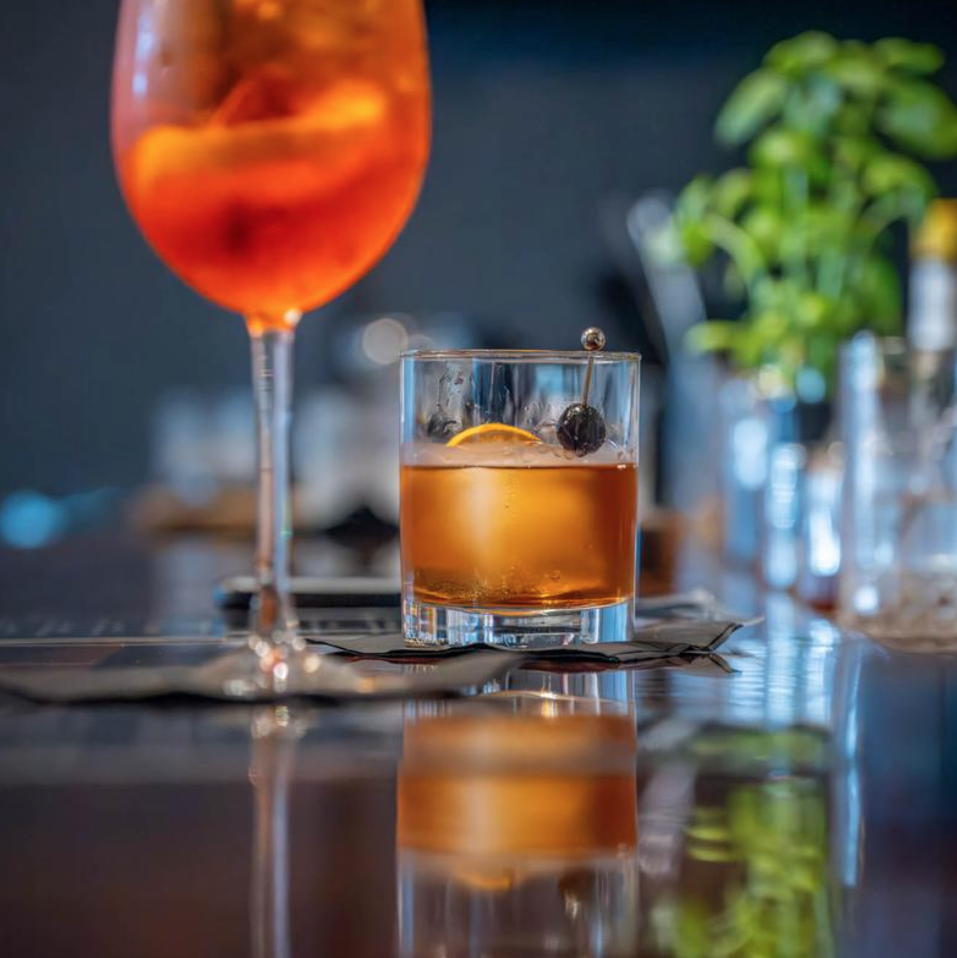 A glass of amber-colored cocktail with a lemon slice and cherry garnish, placed on a napkin on a reflective dark table. Blurred drink in the background with a large ice cube, lemon wedge, and cherry, along with potted plants and glass bottles in the